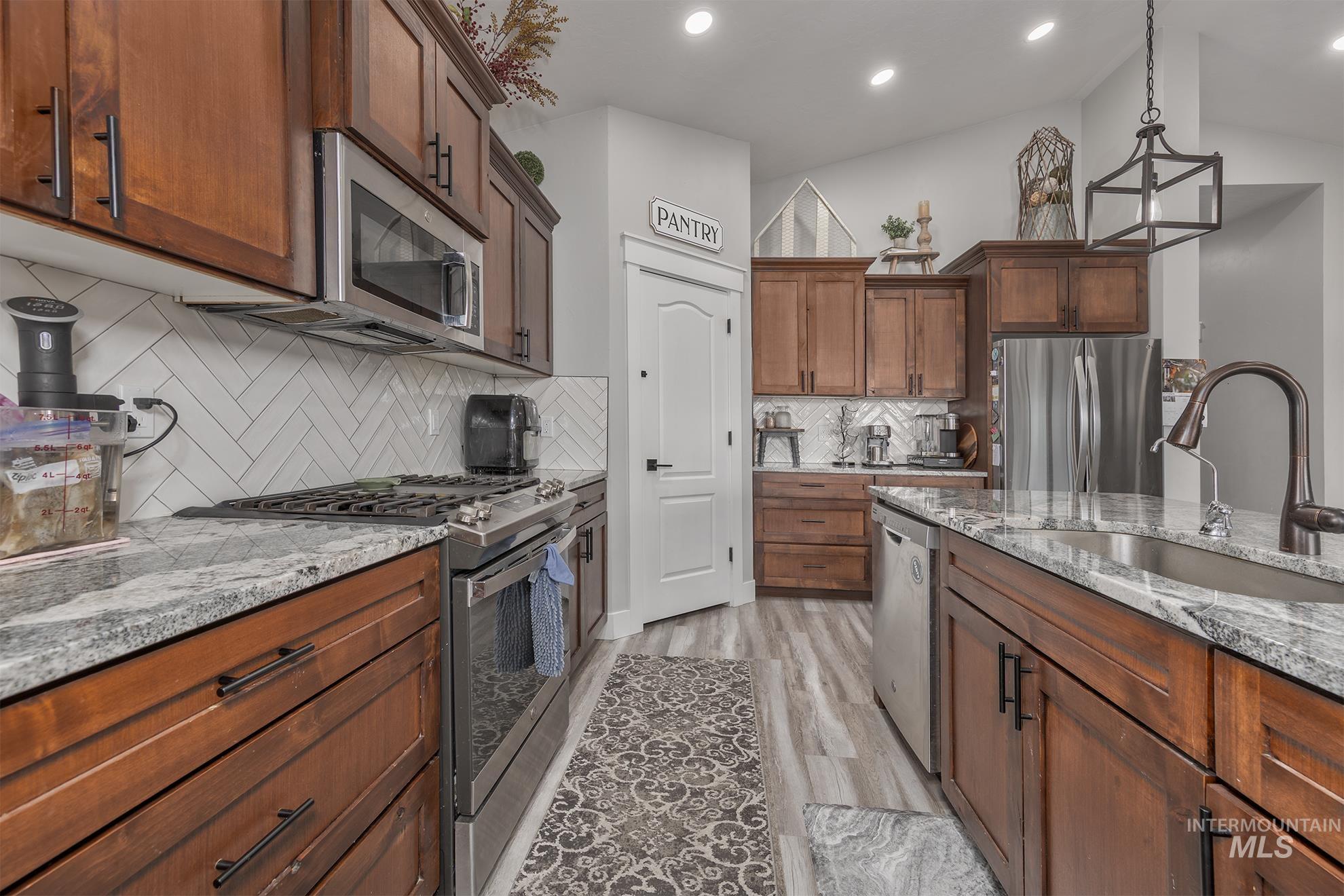 Kitchen featuring appliances with stainless steel finishes, hanging light fixtures, tasteful backsplash, light stone countertops, and brown cabinets