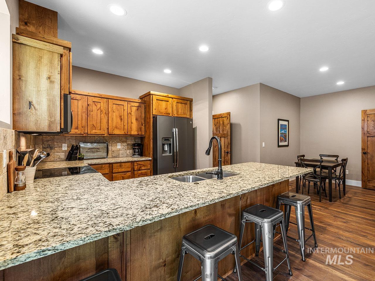 Kitchen with brown cabinets, stainless steel fridge, backsplash, dark wood-style flooring, and light stone counters