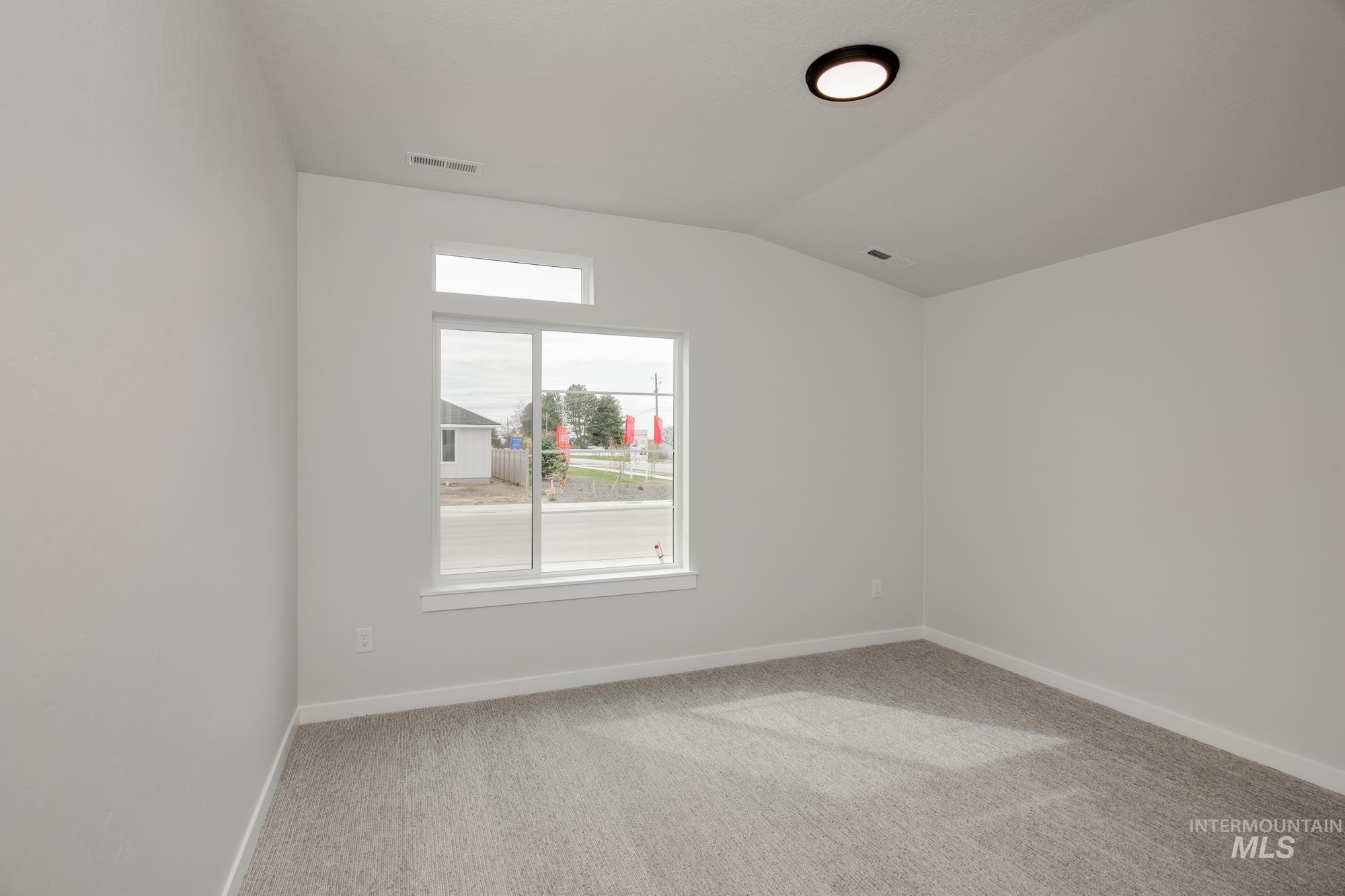 Light-filled room featuring a large double-pane window with transom, neutral carpeting, crisp white walls, a vaulted ceiling, and a flush-mount light fixture