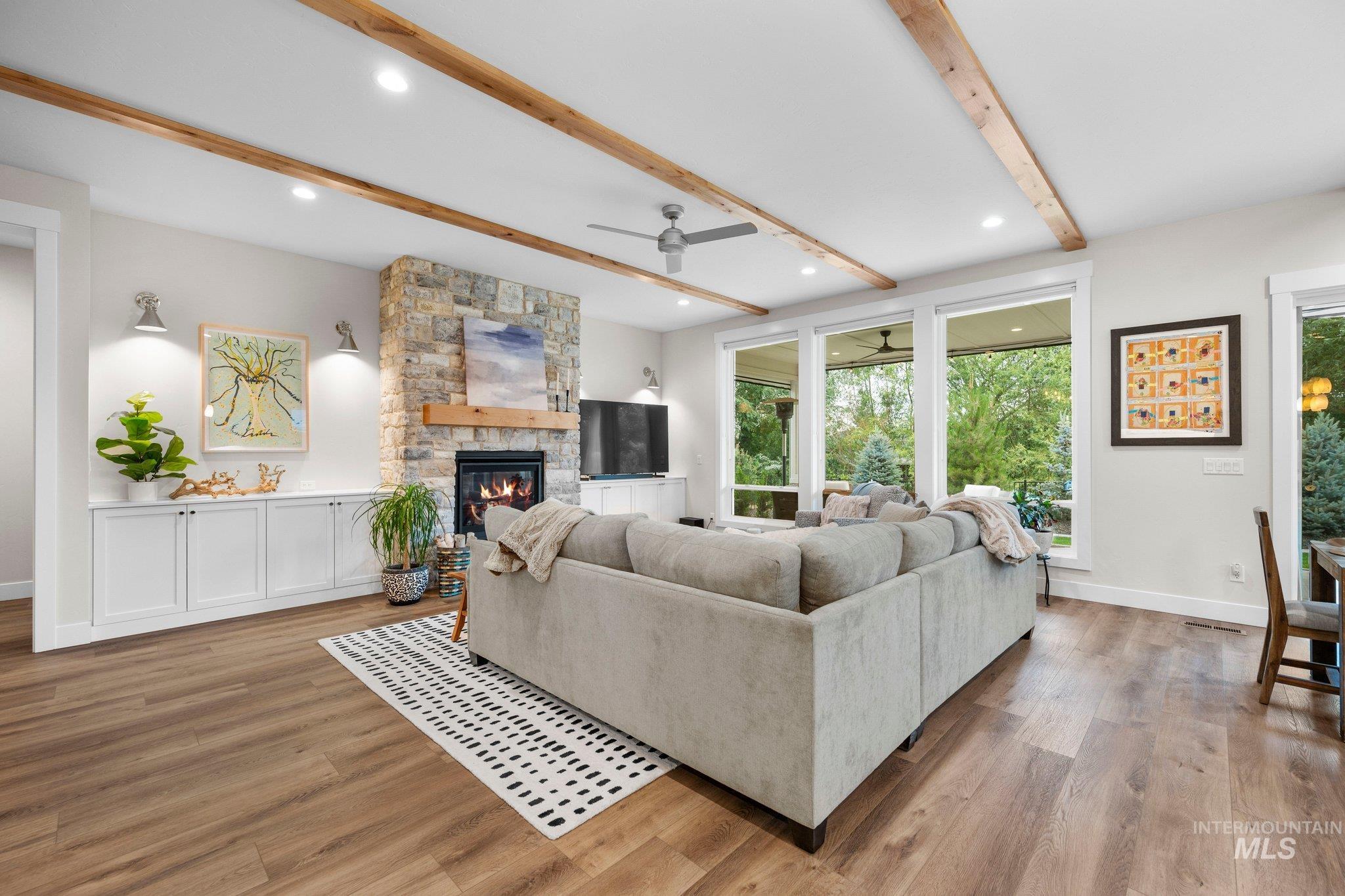 Living room with a stone fireplace, a ceiling fan, light wood-style floors, beam ceiling, and plenty of natural light