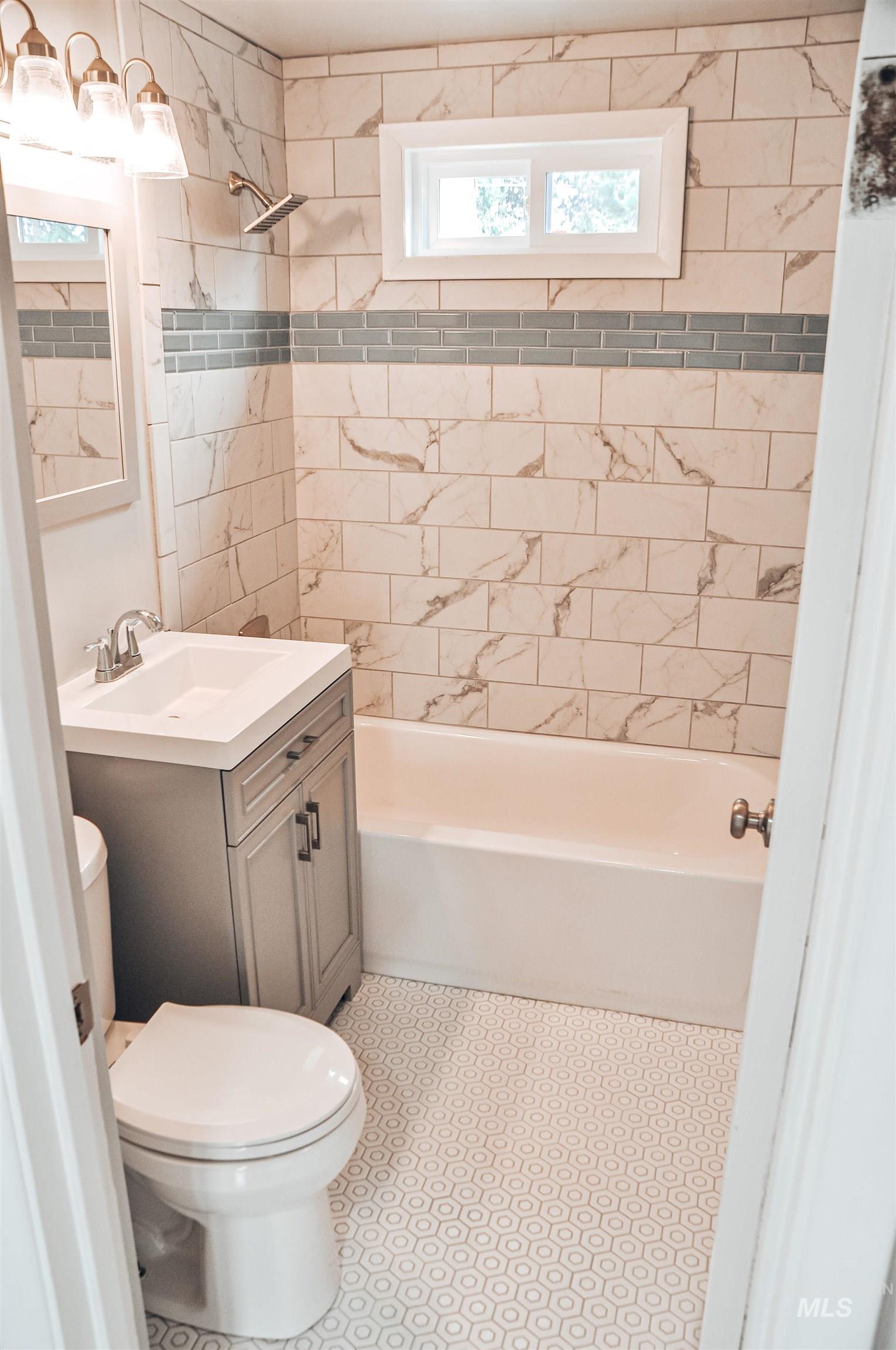 Bathroom featuring full-height tiled walls with a decorative accent band, a built-in bathtub, a window above the tub, a vanity with an integrated sink, and patterned hexagonal floor tiles
