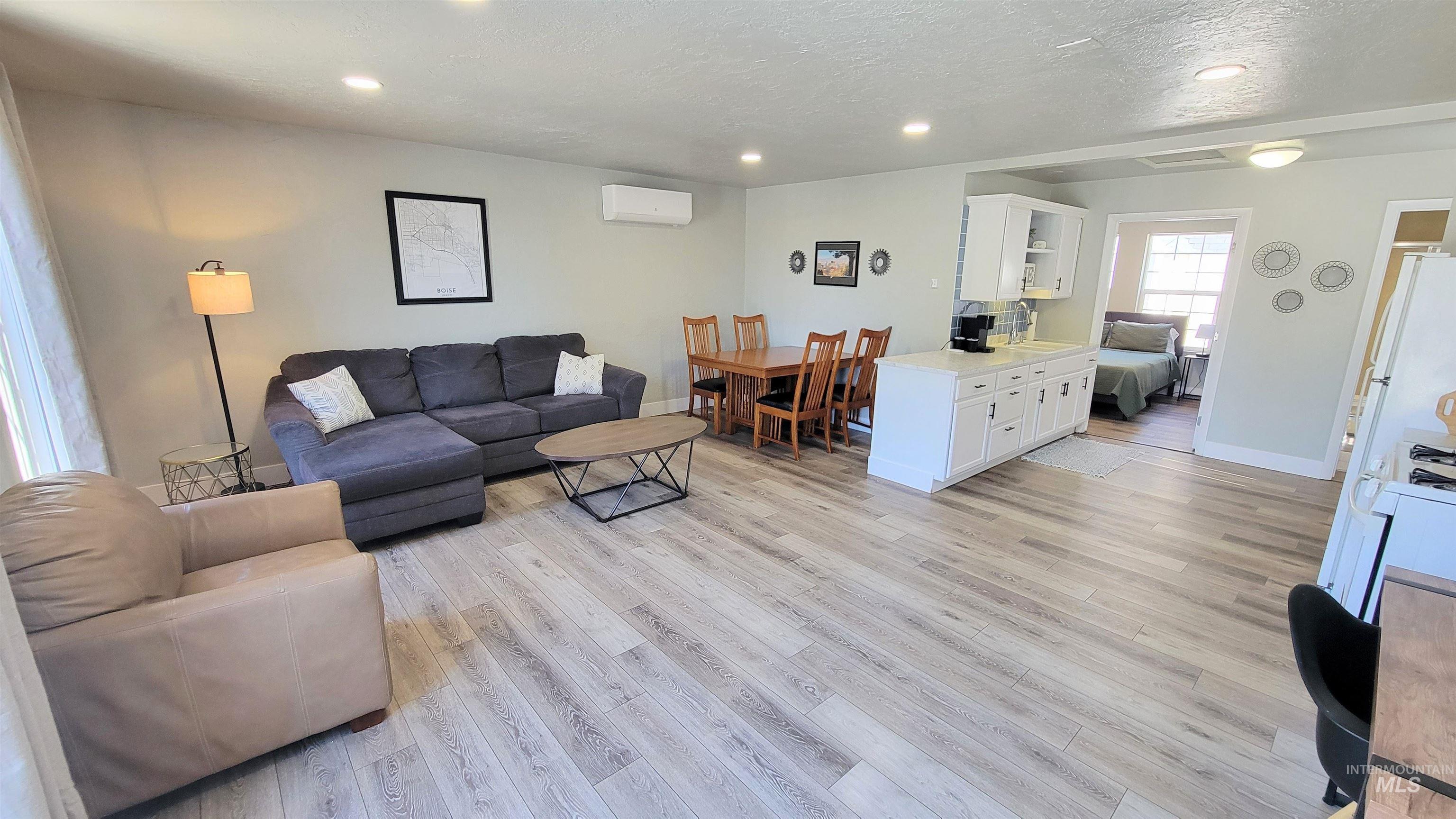 Open-concept living area featuring light wood-finish flooring, recessed ceiling lighting, a dedicated dining space with a wooden table, a kitchen island with white cabinetry, and a mini-split climate control system