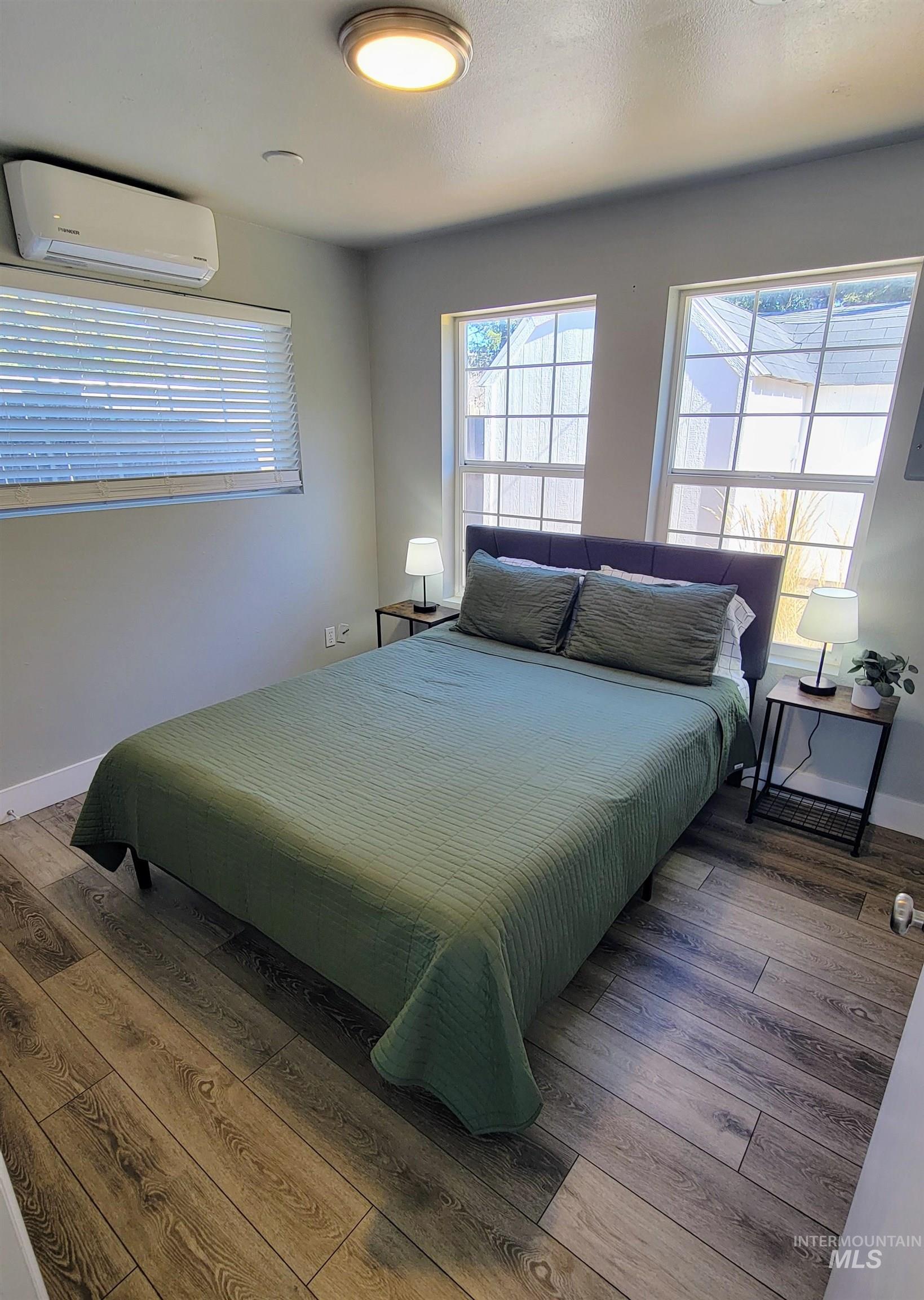Room featuring wood-finish flooring, multiple windows with blinds, a ceiling-mounted light fixture, and a wall-mounted mini-split HVAC unit