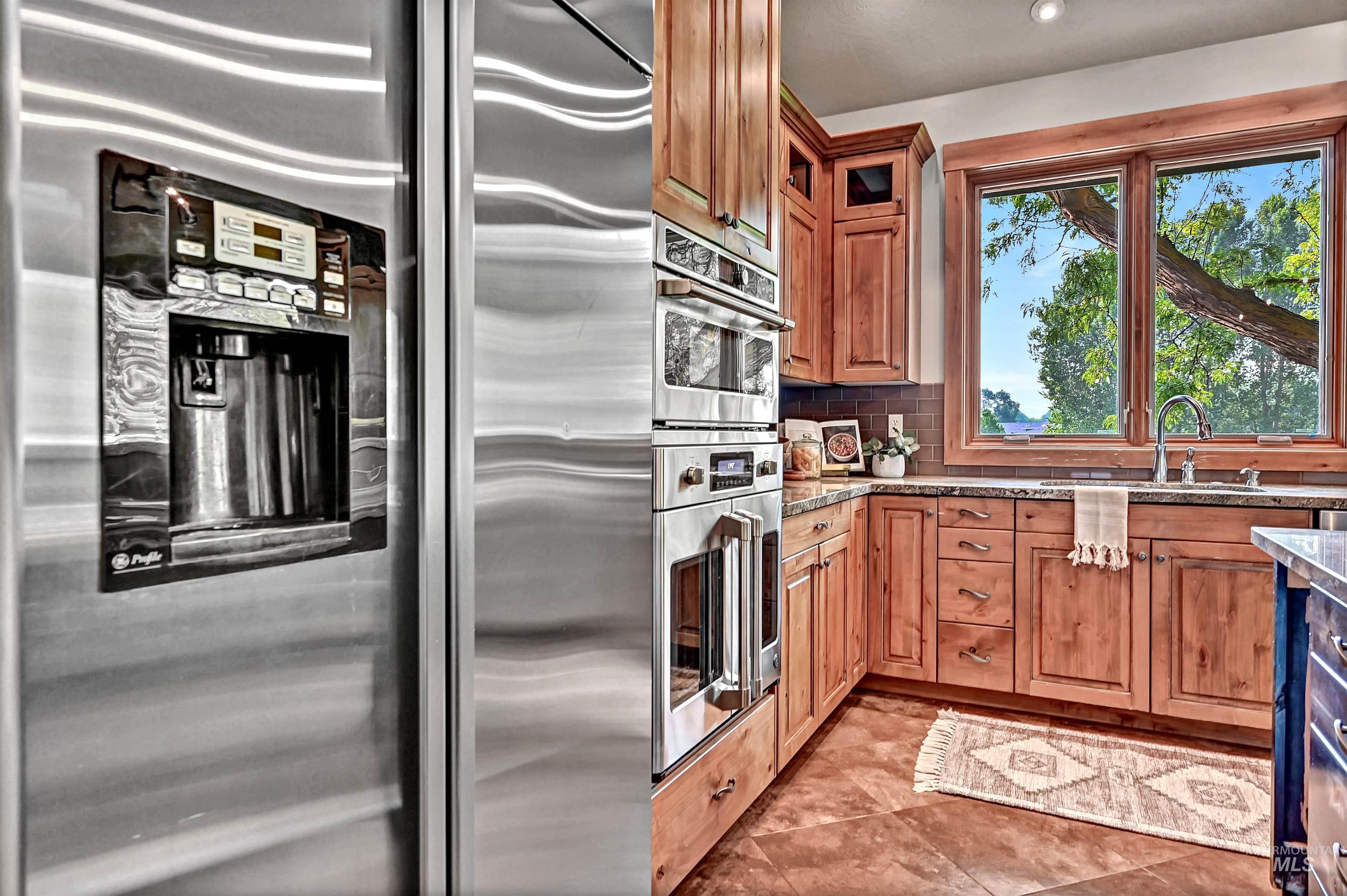 Kitchen featuring stainless steel appliances, decorative backsplash, light stone counters, brown cabinets, and glass insert cabinets