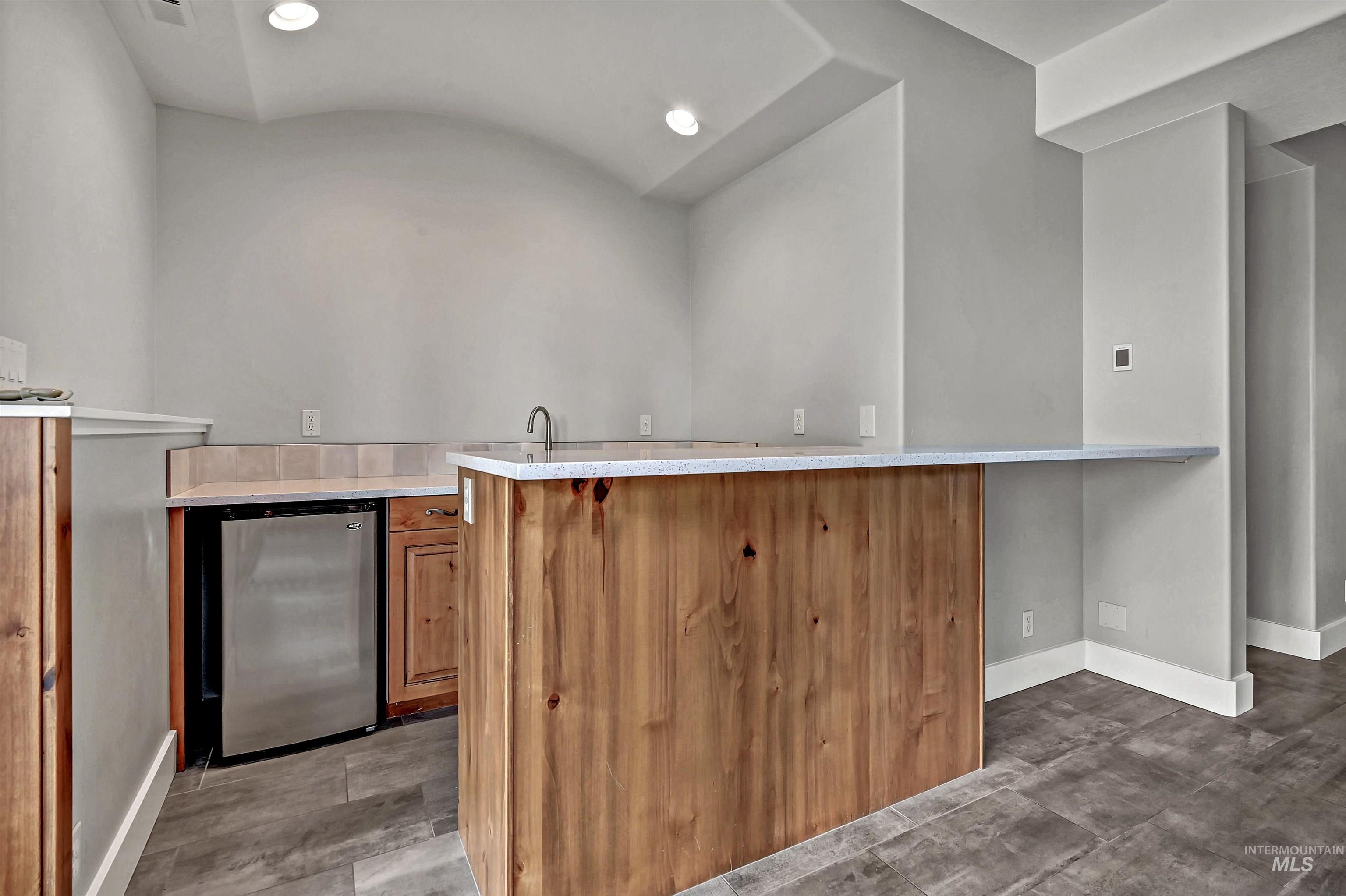 Bar with brown cabinetry, recessed lighting, stainless steel refrigerator, vaulted ceiling, and light stone counters