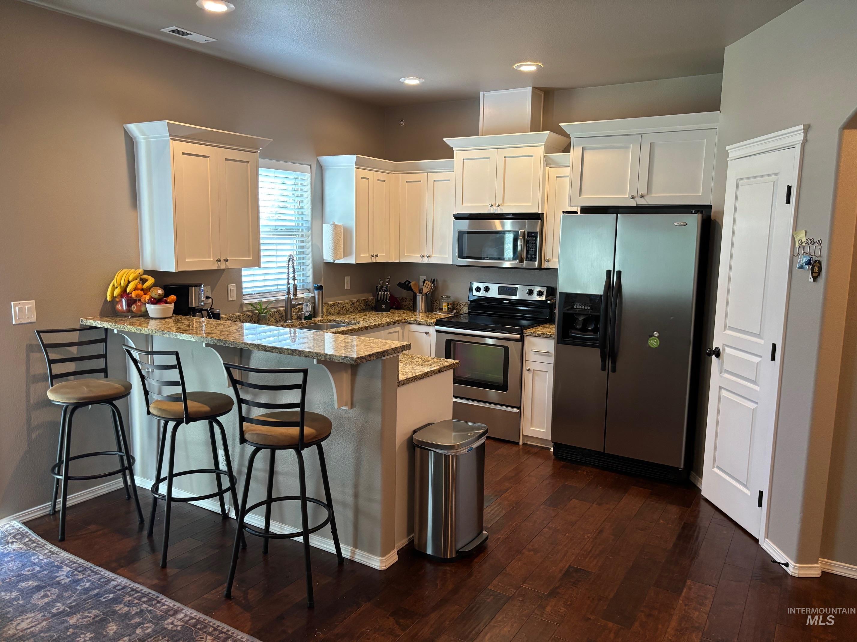 Kitchen with a peninsula, a breakfast bar, stainless steel appliances, light stone countertops, and white cabinets