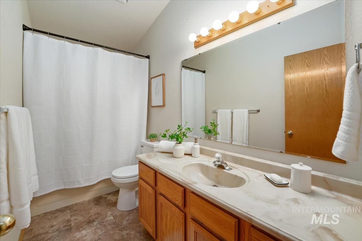 Bathroom featuring a vanity with wood-finish cabinetry and an integrated sink, a large wall-mounted mirror with overhead vanity lighting, a commode, and a shower with a shower curtain