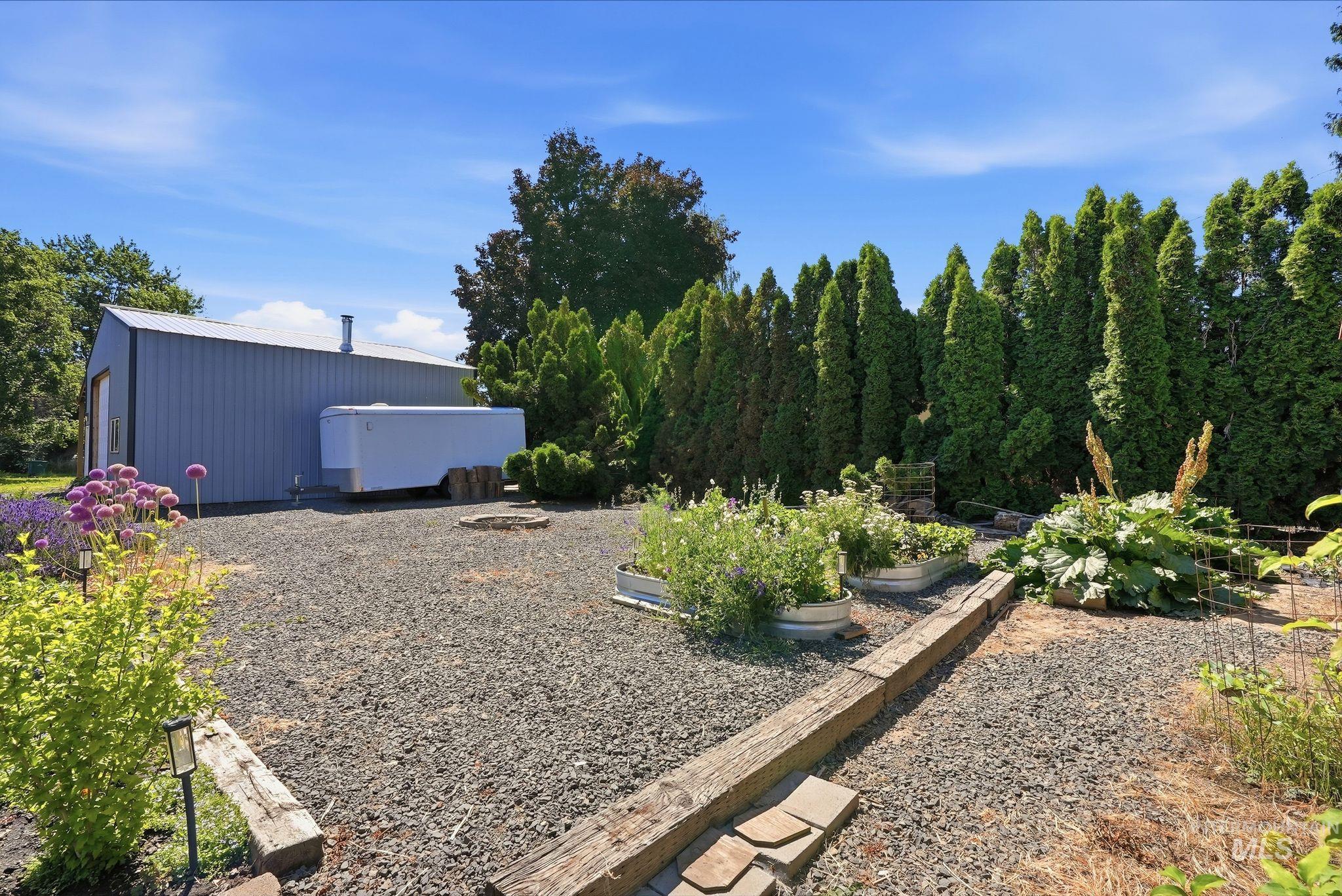 Gravel-covered outdoor space featuring a metal-sided outbuilding with a pitched metal roof, raised garden beds, mature evergreen trees, and integrated landscape lighting