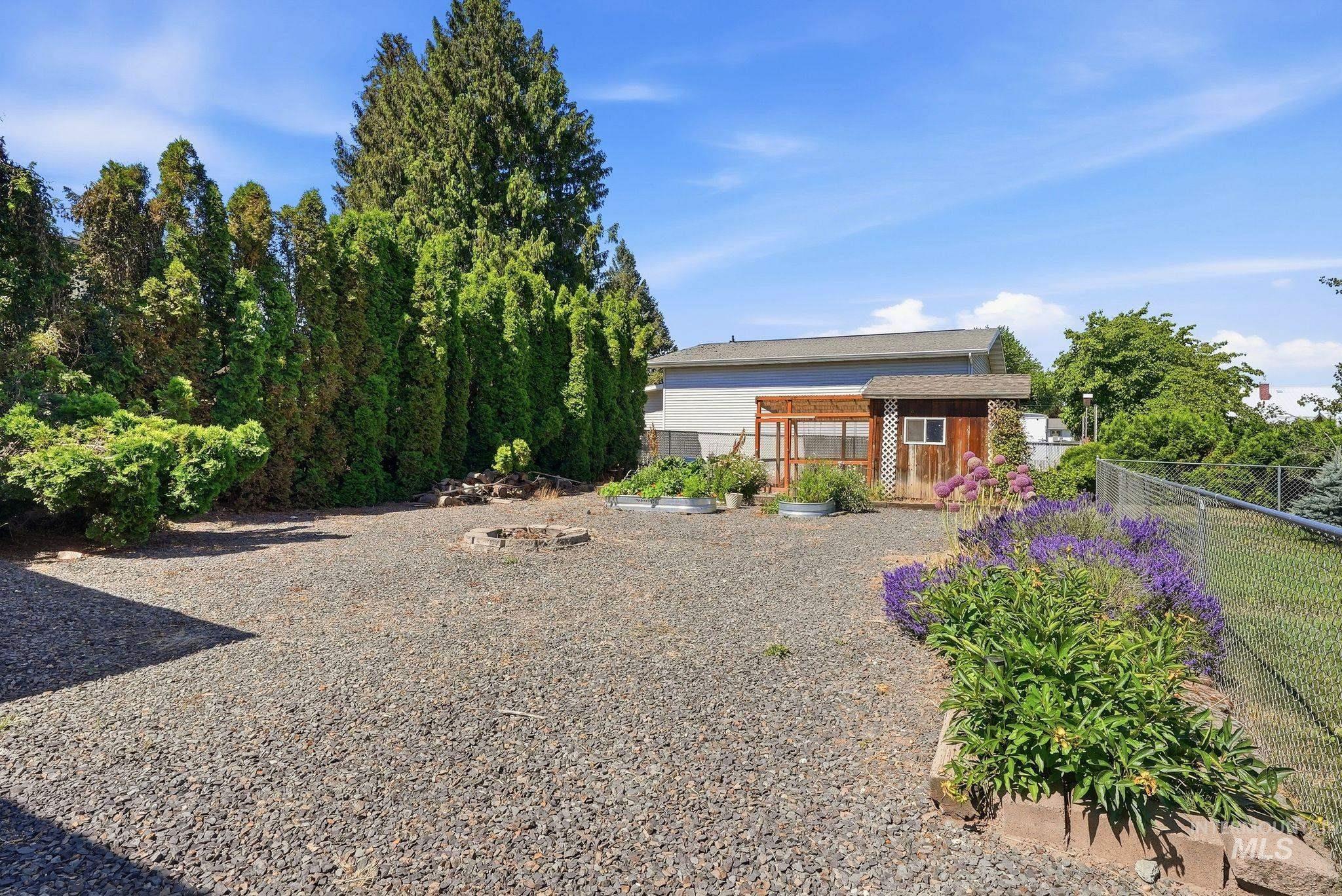 Expansive gravel backyard featuring a built-in fire pit, mature perimeter landscaping, a wood-sided shed with a lattice accent, a fenced property boundary, and a raised garden bed with established plantings