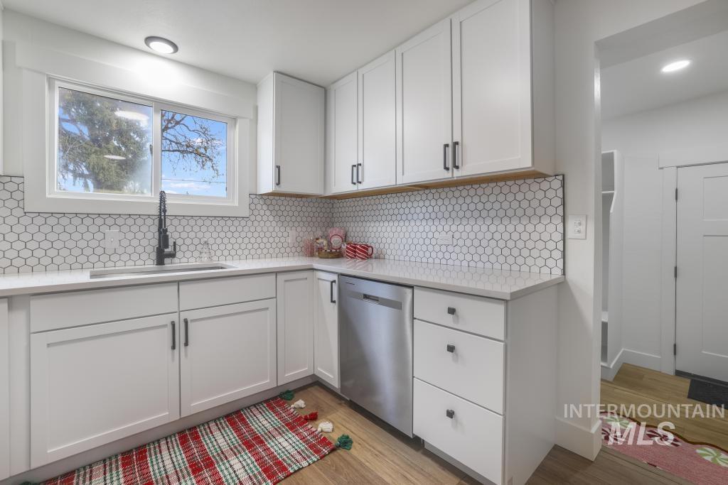 Kitchen with white cabinetry, stainless steel dishwasher, light stone countertops, light wood-style flooring, and decorative backsplash