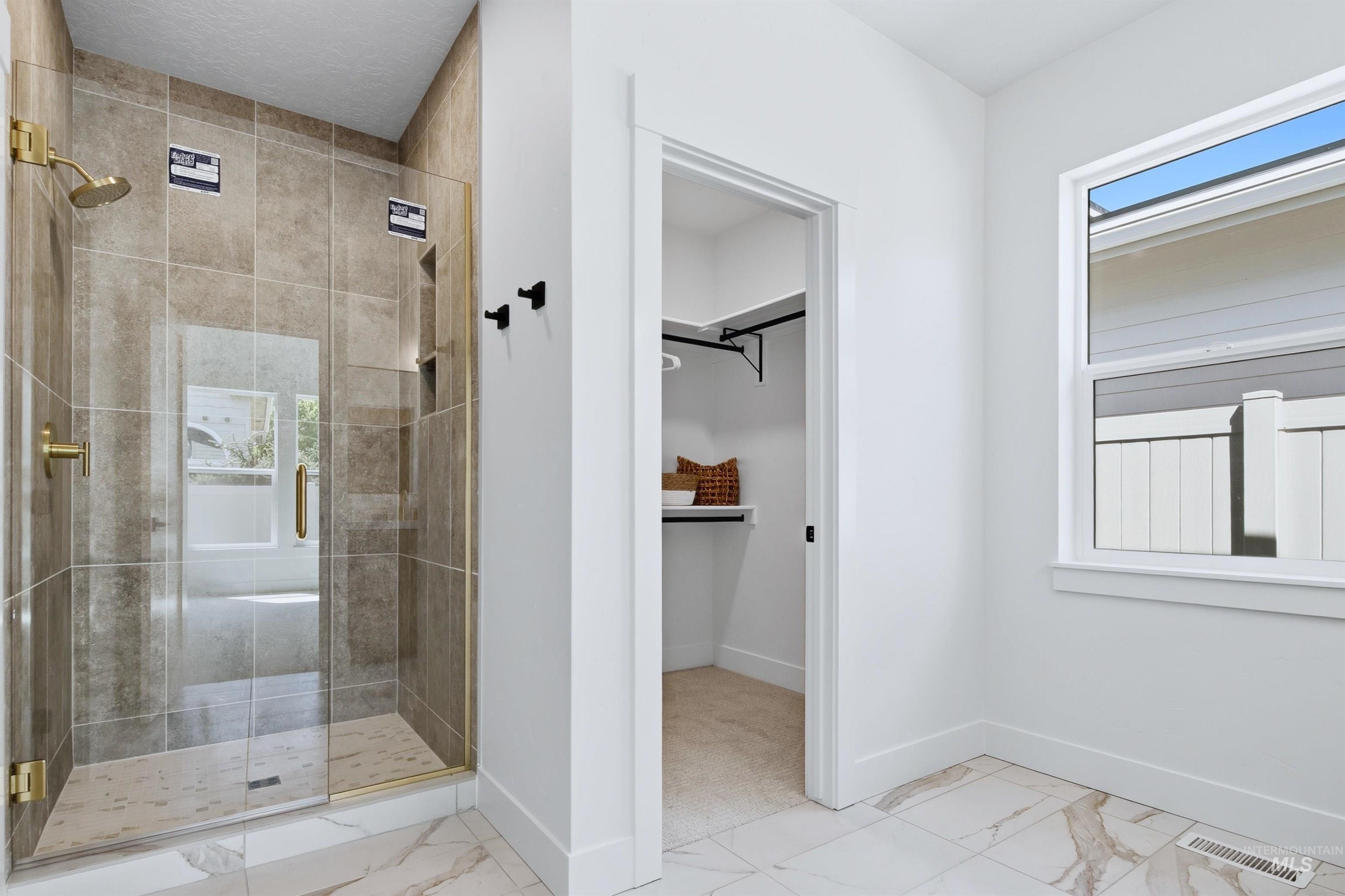 Spacious bathroom featuring a glass-enclosed shower with gold-finish fixtures and brown-toned tile surround, white wall paint, and white marble-look floor tiling