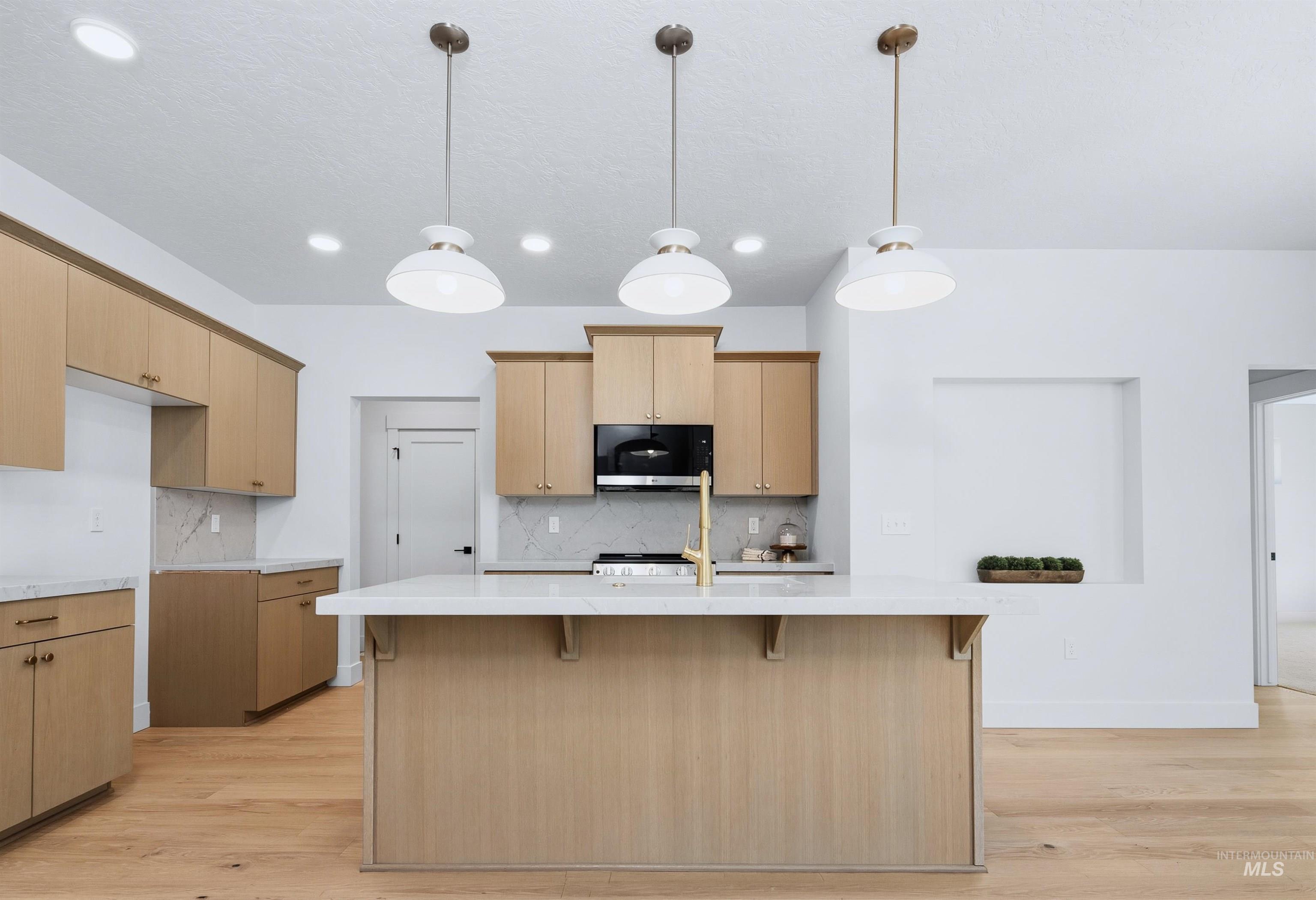Kitchen featuring light wood-finish cabinetry, a central island with a white countertop, gold-tone fixtures, a built-in cooktop with a range hood, and three pendant lights