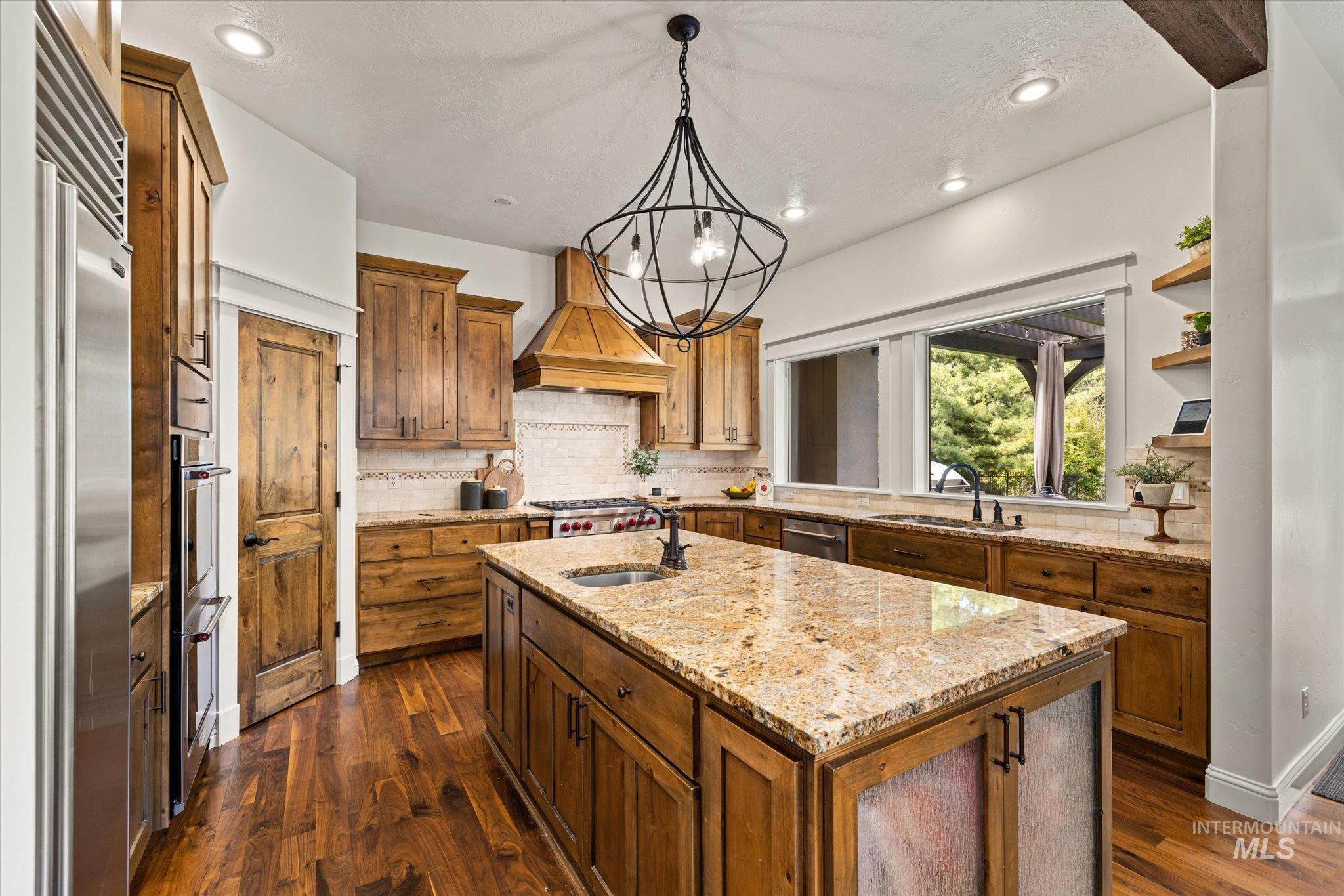 Kitchen with decorative light fixtures, light stone countertops, decorative backsplash, custom exhaust hood, and a kitchen island with sink