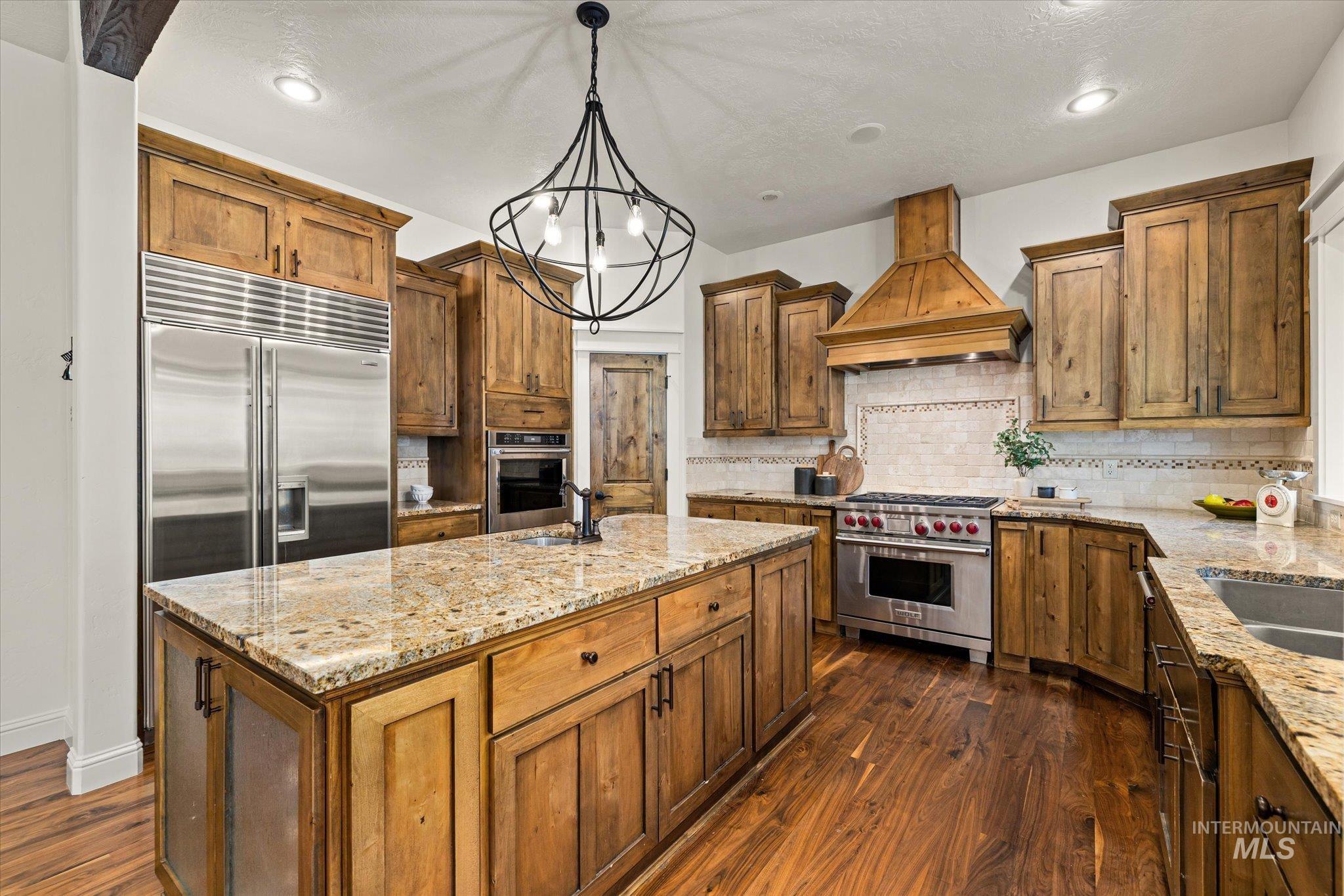 Kitchen with custom exhaust hood, decorative light fixtures, brown cabinetry, light stone countertops, and decorative backsplash