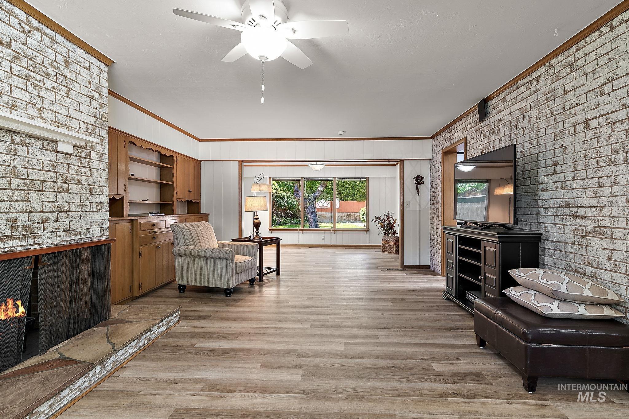 Living area featuring brick wall, crown molding, a brick fireplace, light wood-style flooring, and a ceiling fan