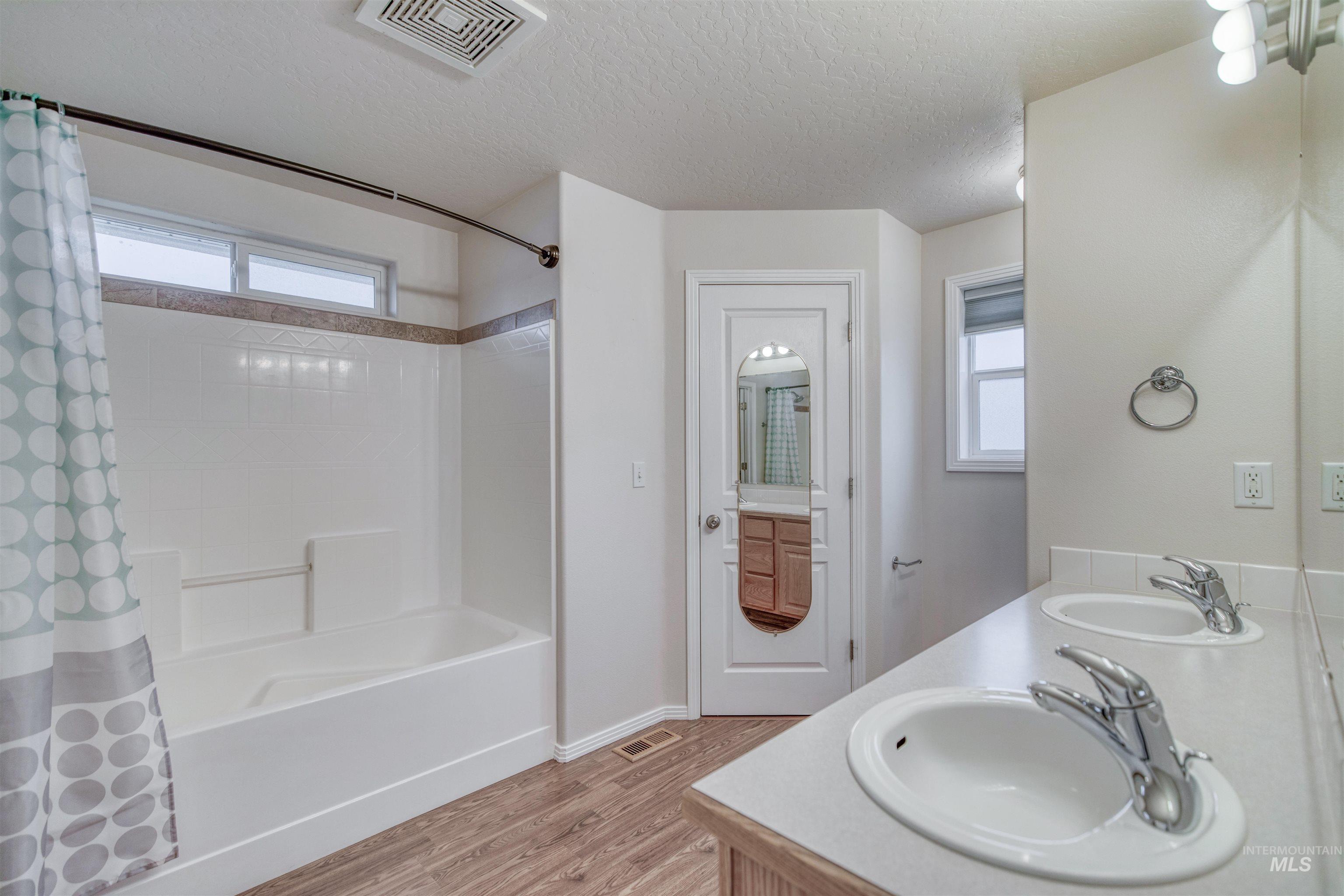 Bathroom featuring healthy amount of natural light, shower / bathtub combination with curtain, light wood-type flooring, double vanity, and a textured ceiling