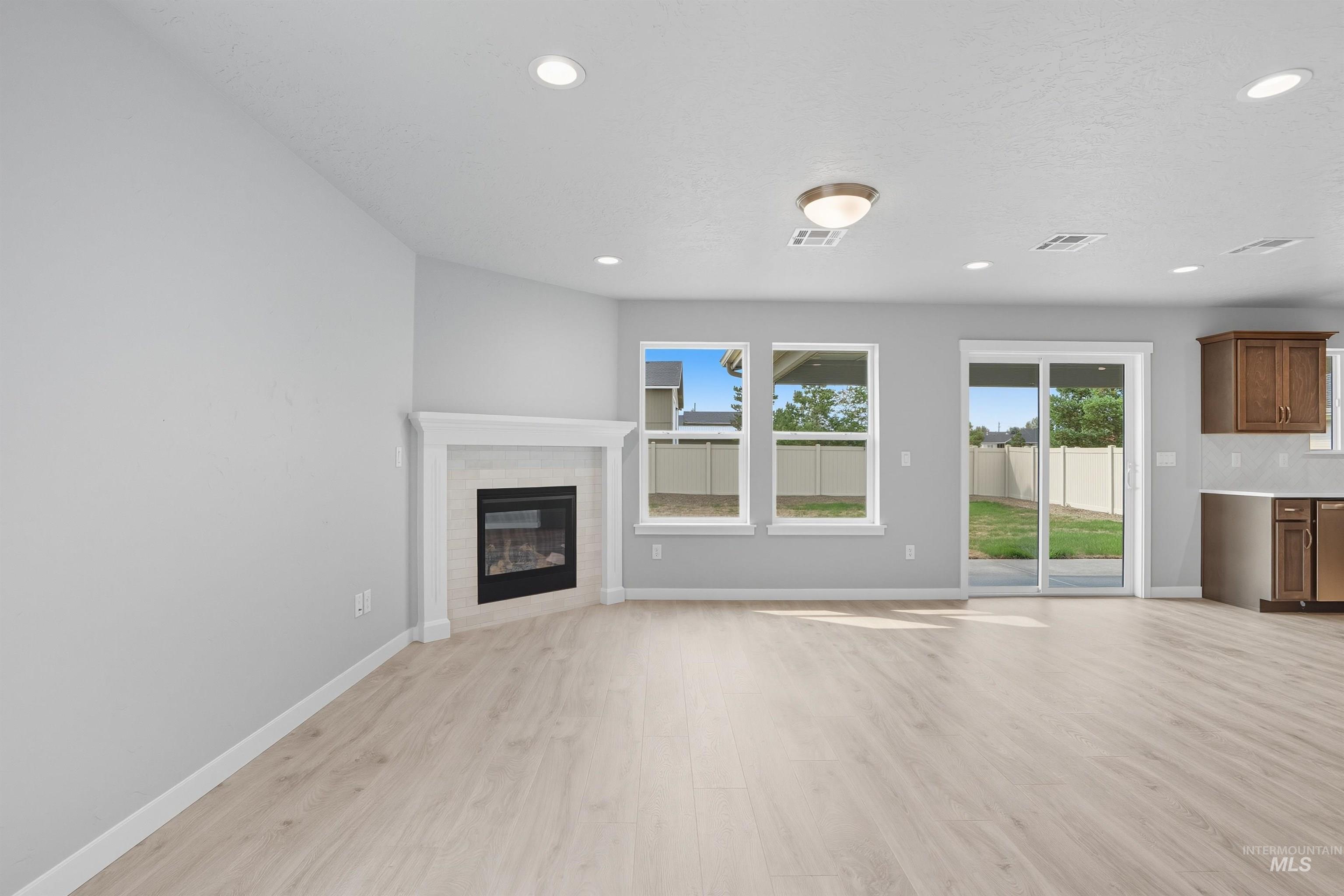 Open-concept living area featuring a corner fireplace with white mantel, light wood-finish flooring, recessed lighting, and a sliding glass door leading to the backyard