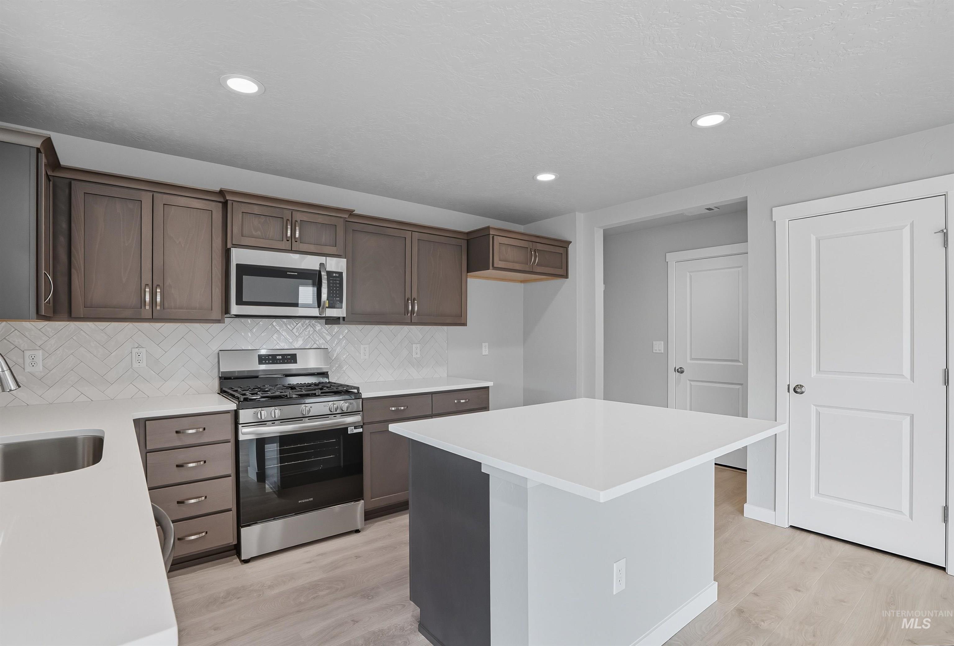 Contemporary kitchen featuring wood-finish cabinetry, stainless steel appliances, a herringbone tile backsplash, light-toned countertops, and a central island