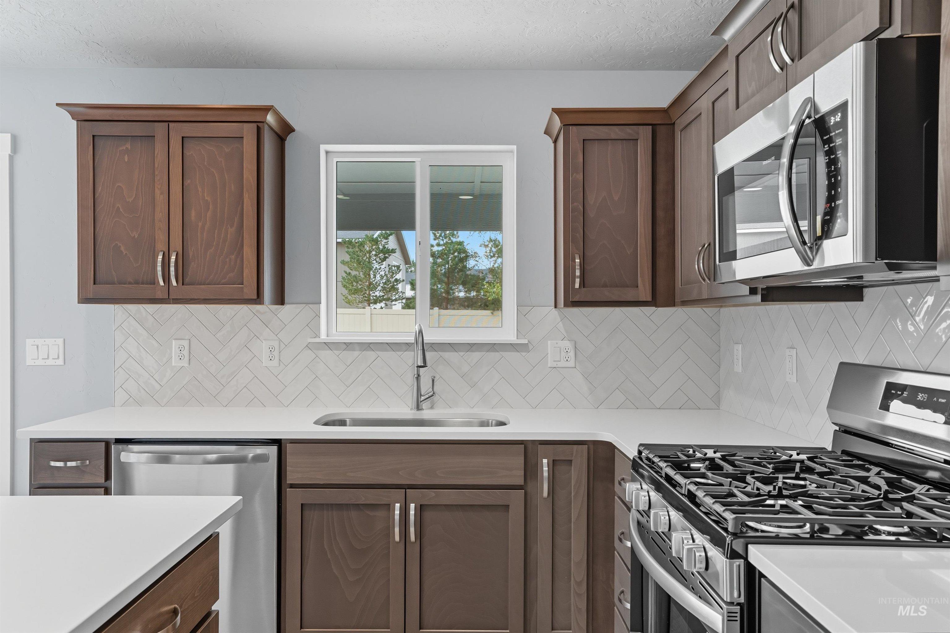 Kitchen featuring rich wood cabinetry, stainless steel appliances, white countertops, a herringbone tile backsplash, and a window above the sink
