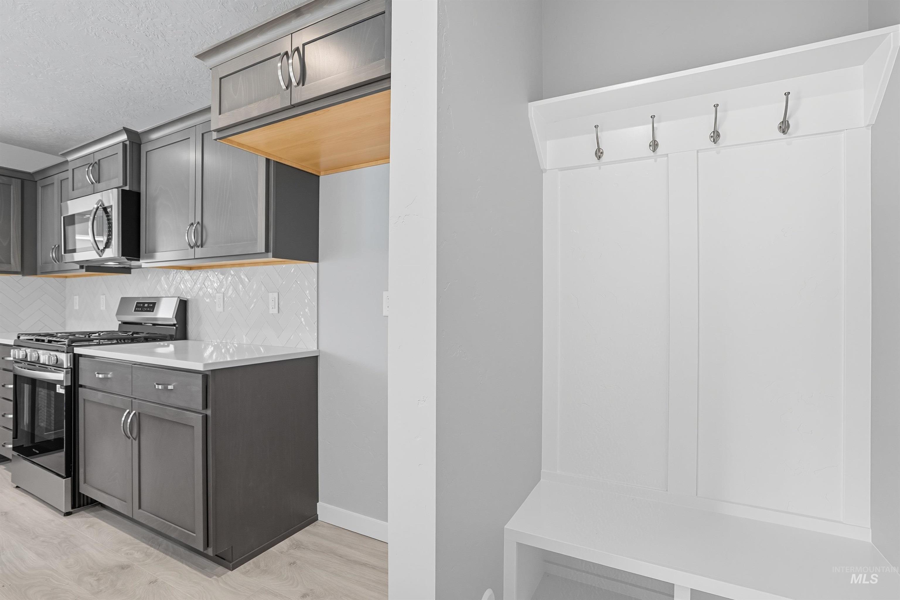 Built-in mudroom bench with coat hooks complementing a contemporary kitchen featuring stainless steel appliances, grey cabinetry, light countertops, and a white geometric tile backsplash