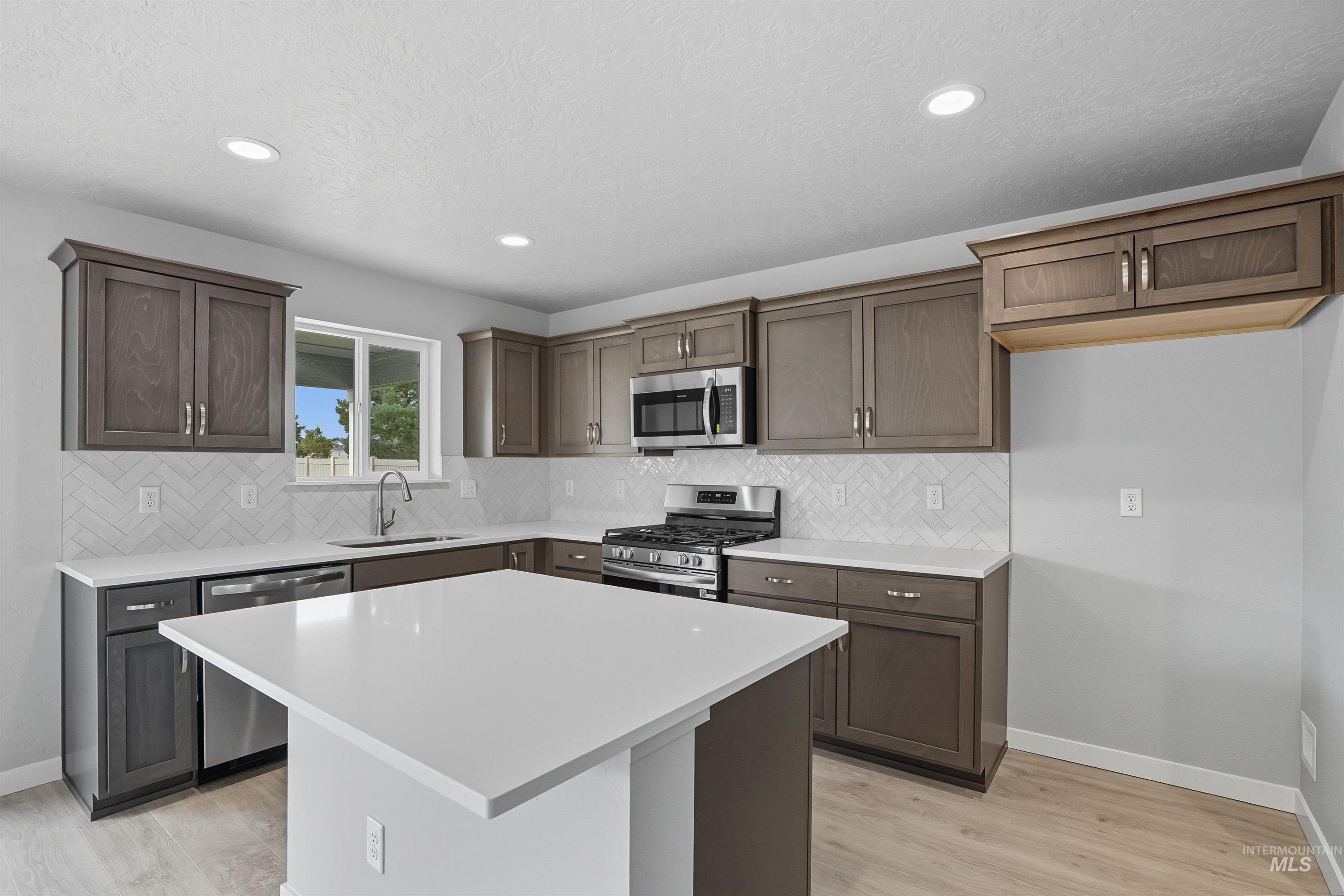 Modern kitchen featuring a central island with a white countertop, wood-finish flooring, recessed lighting, stainless steel appliances, and a white herringbone tile backsplash