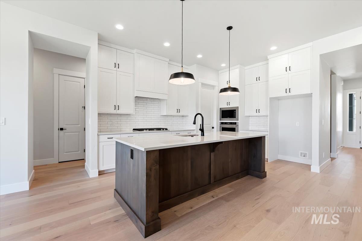 Kitchen featuring dark brown cabinetry, decorative backsplash, white cabinets, light wood-style floors, and an island with sink