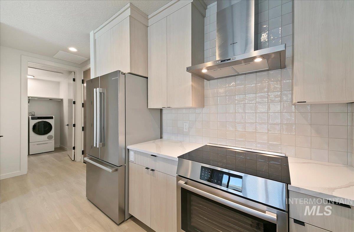 Kitchen with wall chimney exhaust hood, appliances with stainless steel finishes, light stone counters, washer / dryer, and light brown cabinets