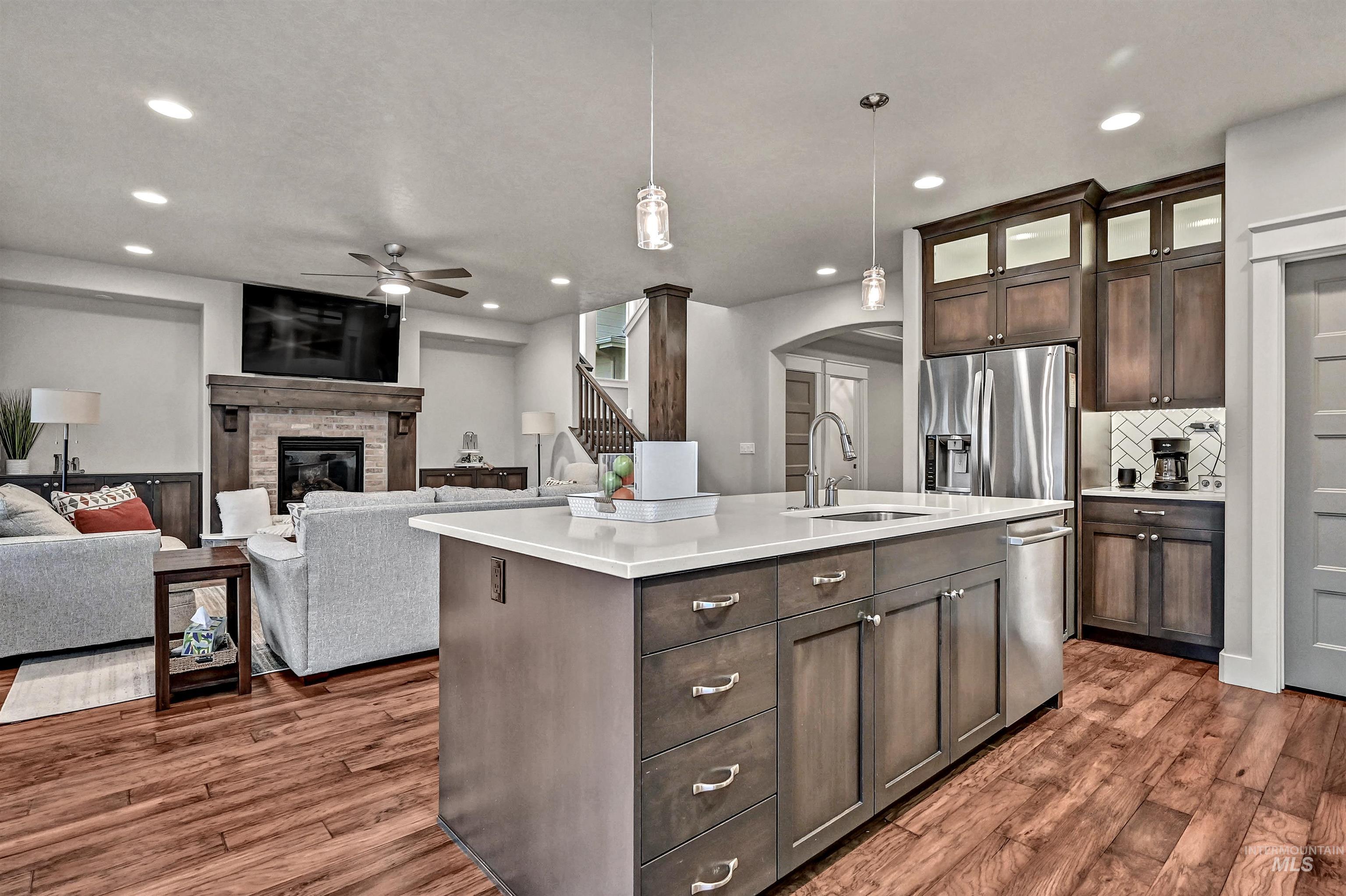 Kitchen featuring decorative light-colored tile backsplash, dark cabinets, appliances with stainless steel finishes, exhaust hood above the gas range, white quartz counters with a center island with a sink, and decorative light fixtures