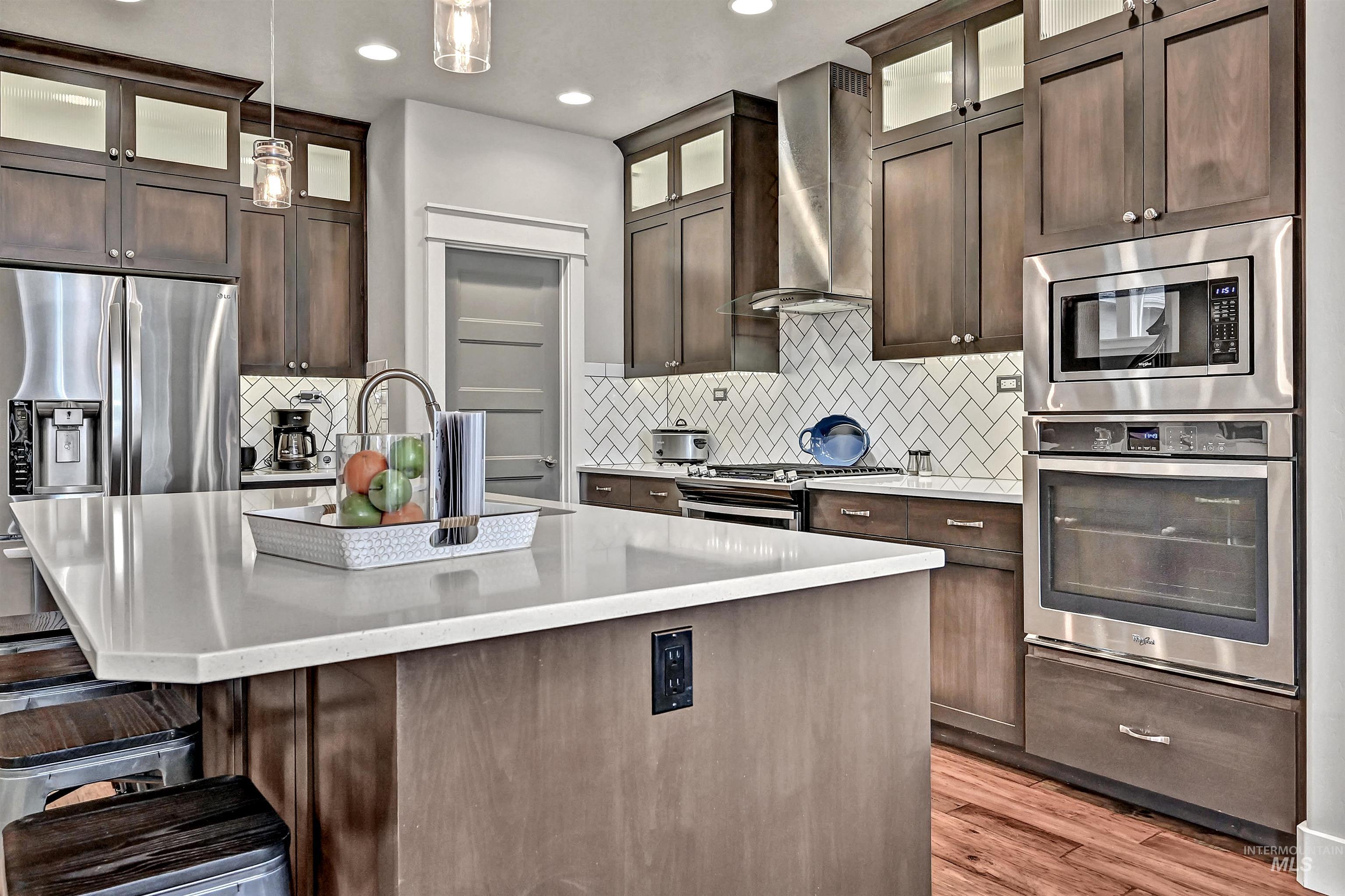 Kitchen featuring decorative light-colored tile backsplash, dark cabinets, appliances with stainless steel finishes, exhaust hood above the gas range, white quartz counters with a center island with a sink, and decorative light fixtures