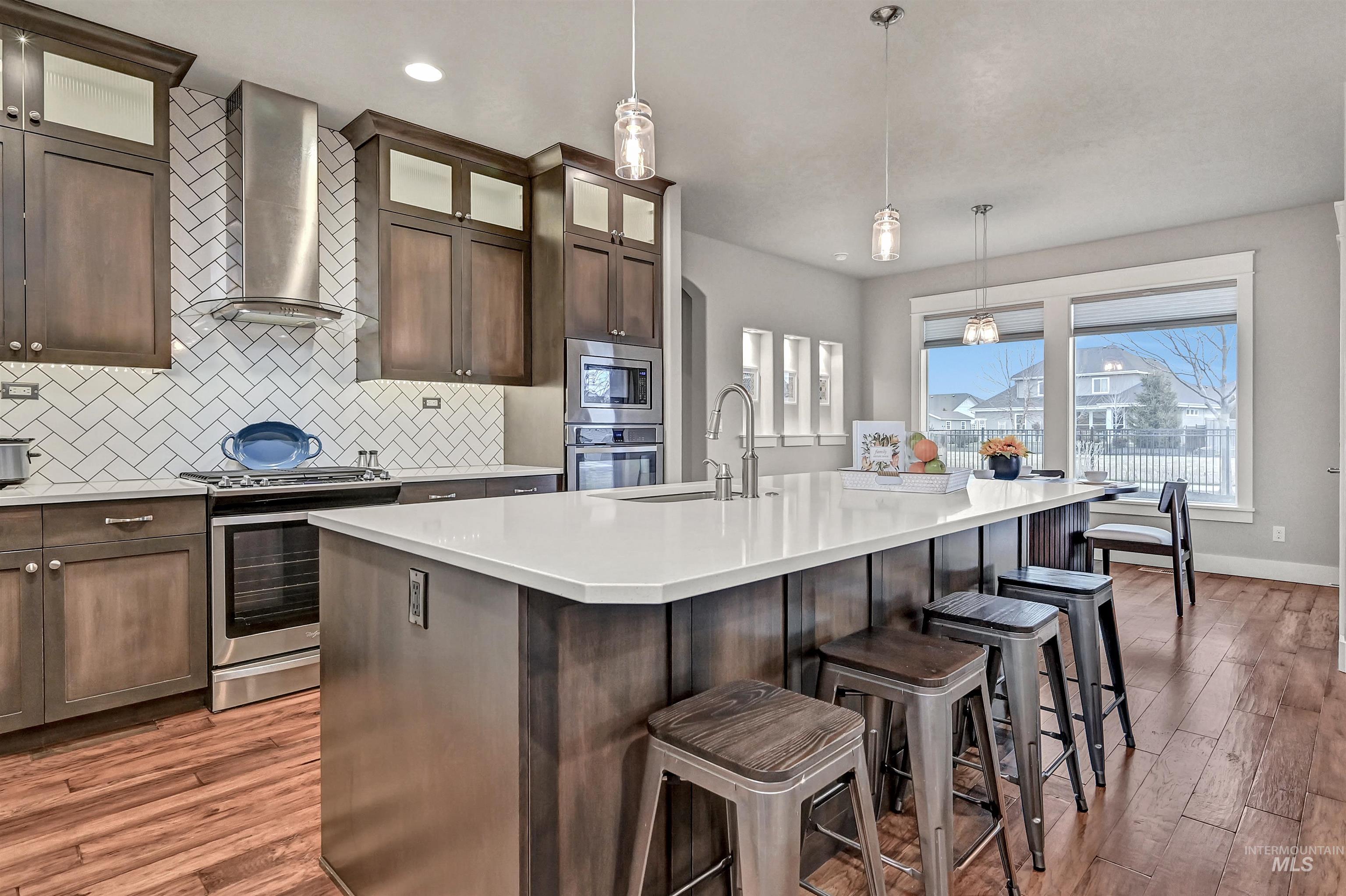 Kitchen featuring decorative light-colored tile backsplash, dark cabinets, appliances with stainless steel finishes, exhaust hood above the gas range, white quartz counters with a center island with a sink, and decorative light fixtures