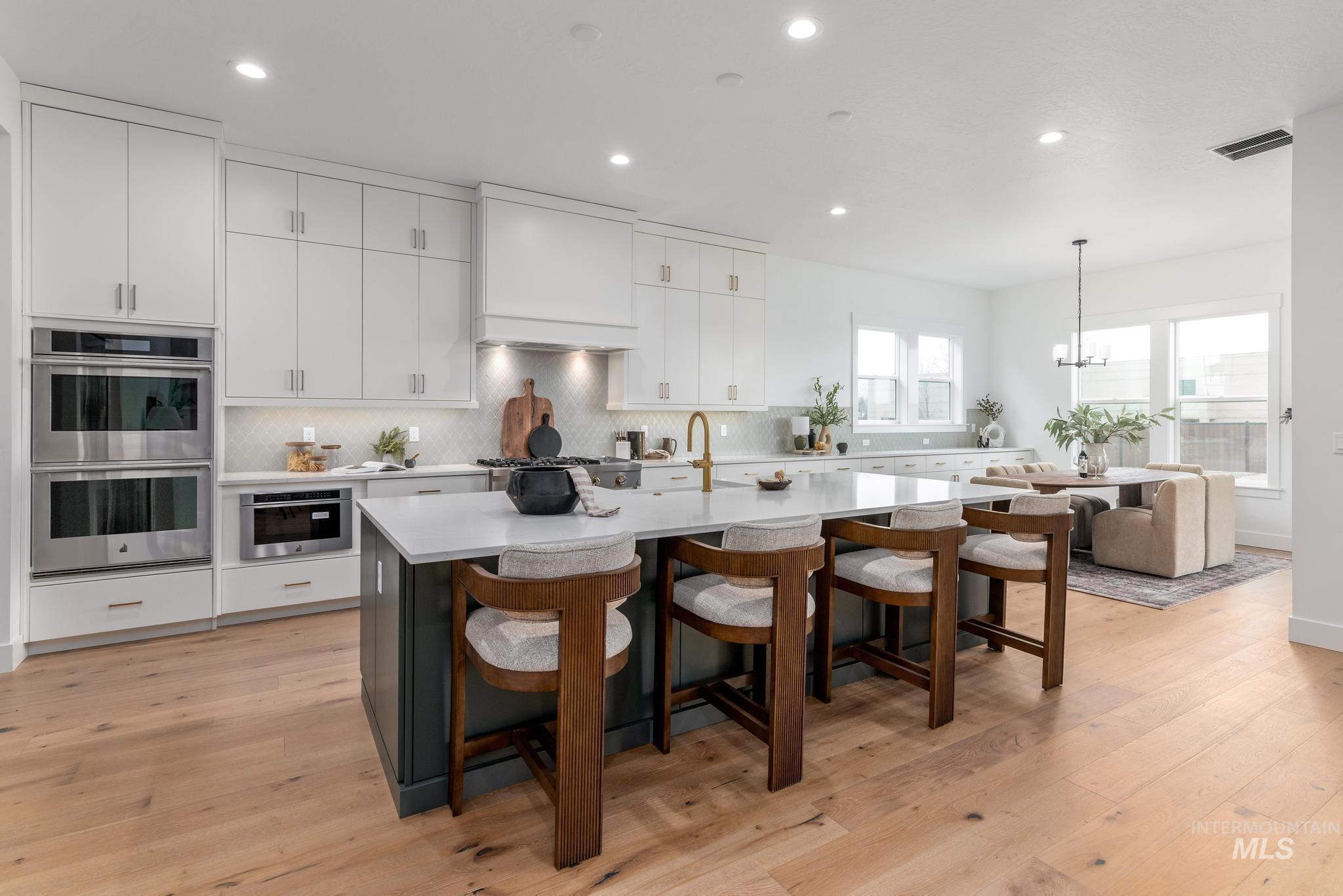 Modern kitchen featuring a large central island with seating, light wood-finish flooring, white upper and lower cabinetry, stainless steel double ovens, and a light-colored backsplash
