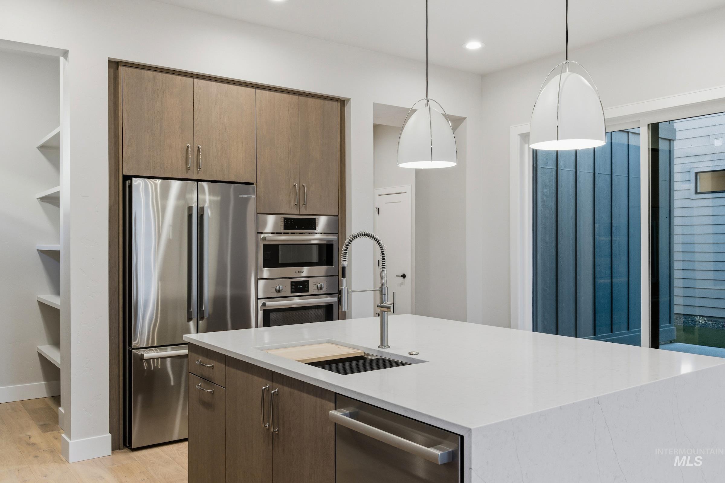 Kitchen with stainless steel appliances, hanging light fixtures, light stone counters, light wood-type flooring, and a center island with sink