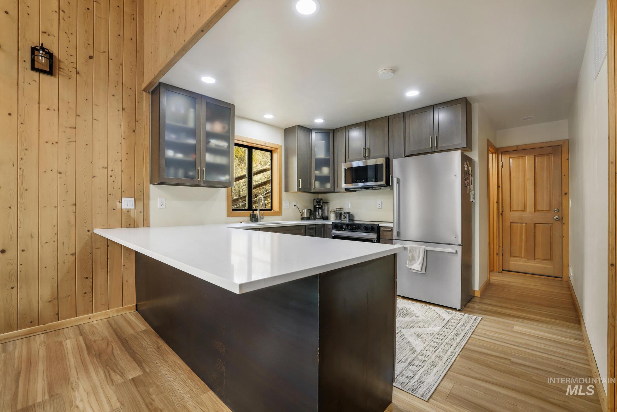 Kitchen with glass insert cabinets, stainless steel appliances, a peninsula, light wood-style flooring, and wood walls