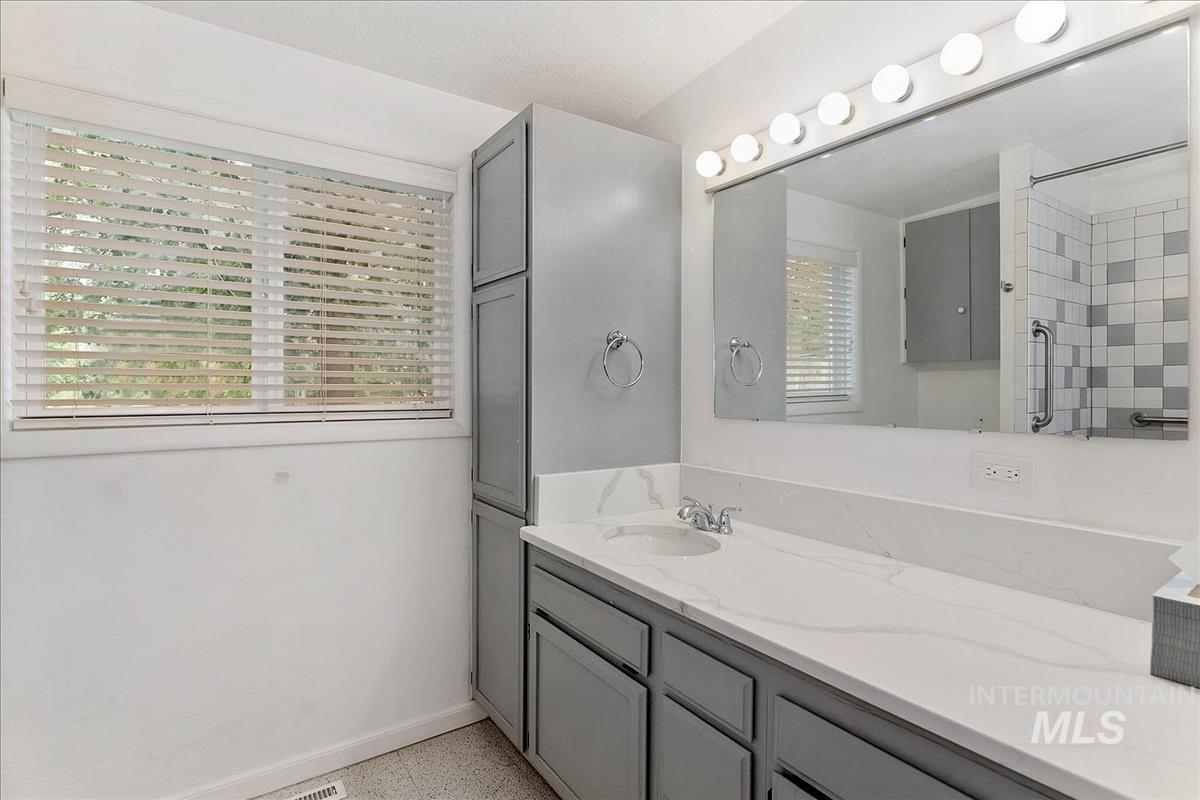 Bathroom featuring an extended vanity with a white countertop and integrated sink, under-cabinet storage, a large wall mirror with vanity lighting, and a window with horizontal blinds