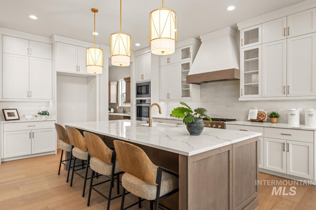 Spacious kitchen featuring an expansive central island with a light-toned countertop, white cabinetry with gold-tone hardware, wood-finish flooring, a built-in range hood with wood accent, and recessed lighting