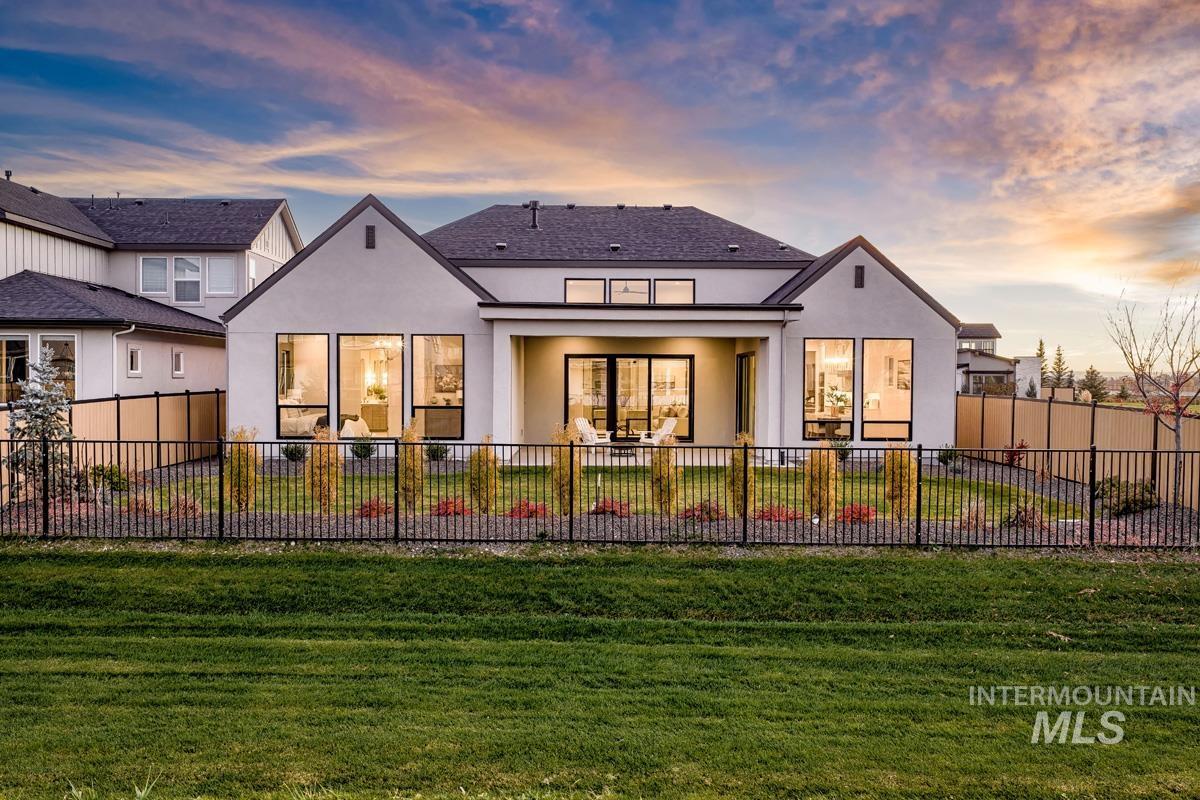 Contemporary rear exterior featuring expansive windows, a covered patio, a dark composite shingle roof, a black wrought iron fence, and established landscaping