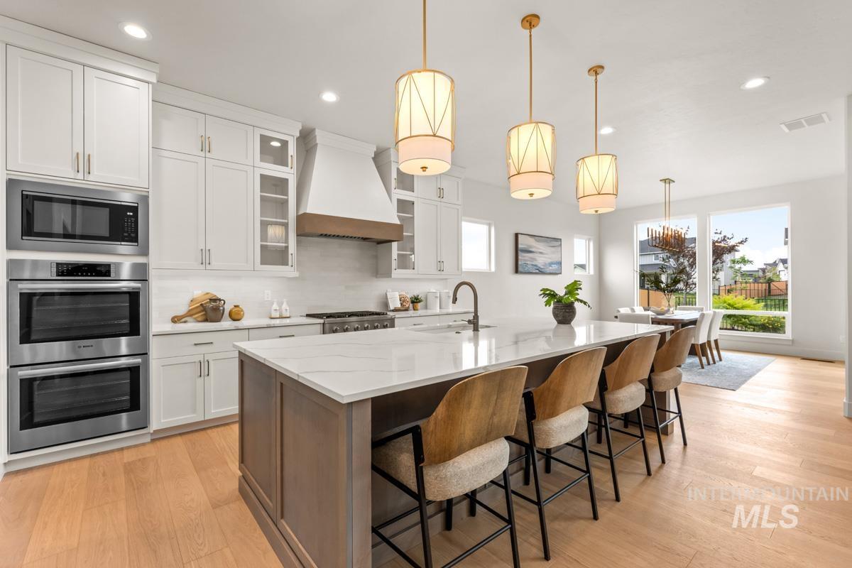 Gourmet kitchen featuring a large central island with seating, stainless steel appliances including double ovens, extensive white cabinetry, and light wood-finish flooring