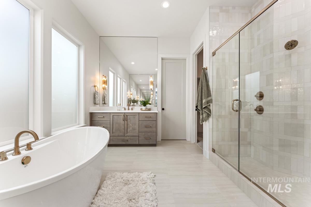 Freestanding soaking tub with brass fixtures, a frameless glass shower, a dual vanity with wood-finish cabinetry, a large wall-mounted mirror, and frosted windows
