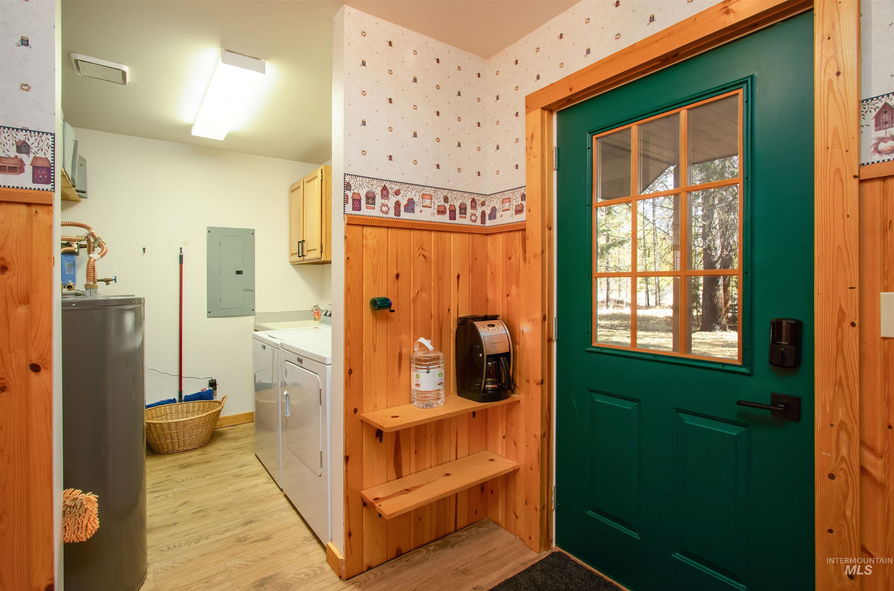 Doorway to outside featuring water heater, wood finished floors, separate washer and dryer, electric panel, and wallpapered walls