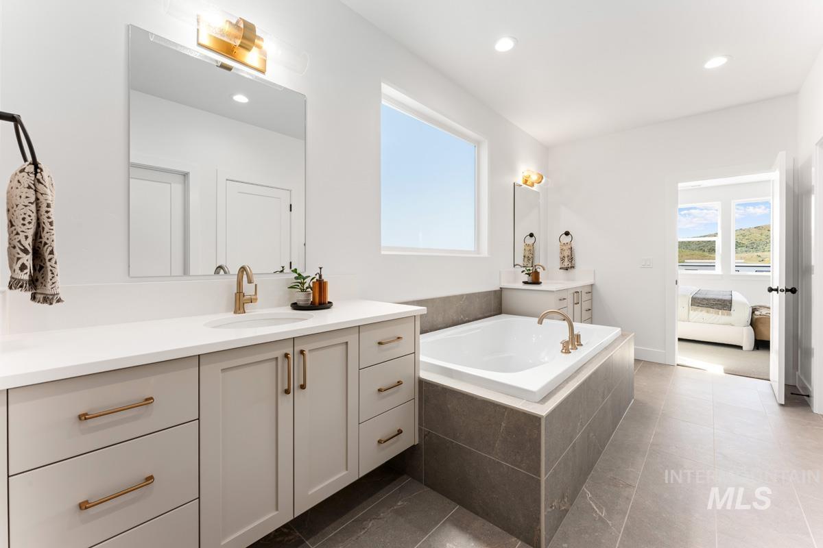 Spacious bathroom featuring a built-in soaking tub with tiled surround, a large window, and a dual vanity with light-toned cabinetry and gold-tone hardware