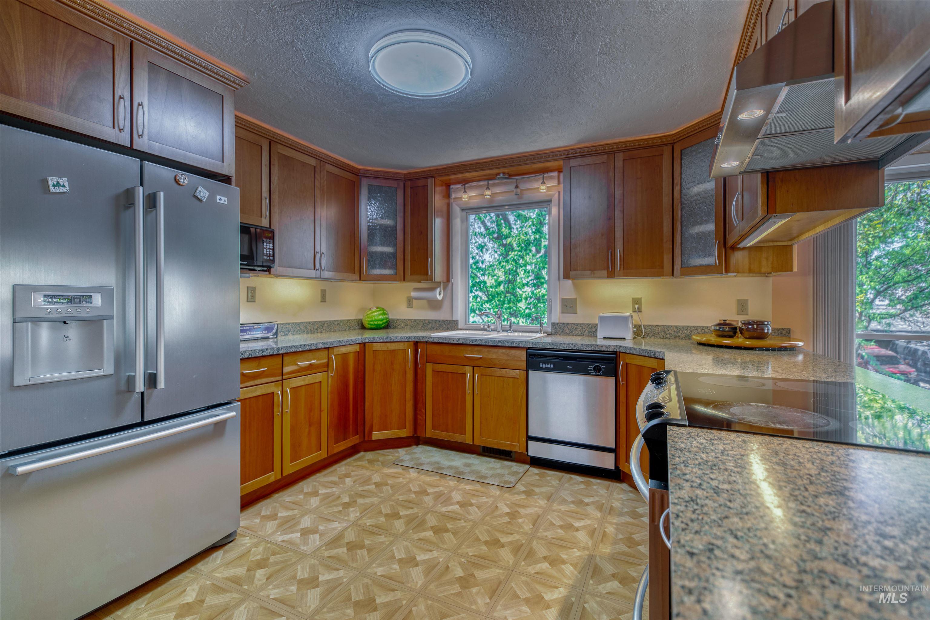 Kitchen featuring appliances with stainless steel finishes, a textured ceiling, glass insert cabinets, under cabinet range hood, and brown cabinets