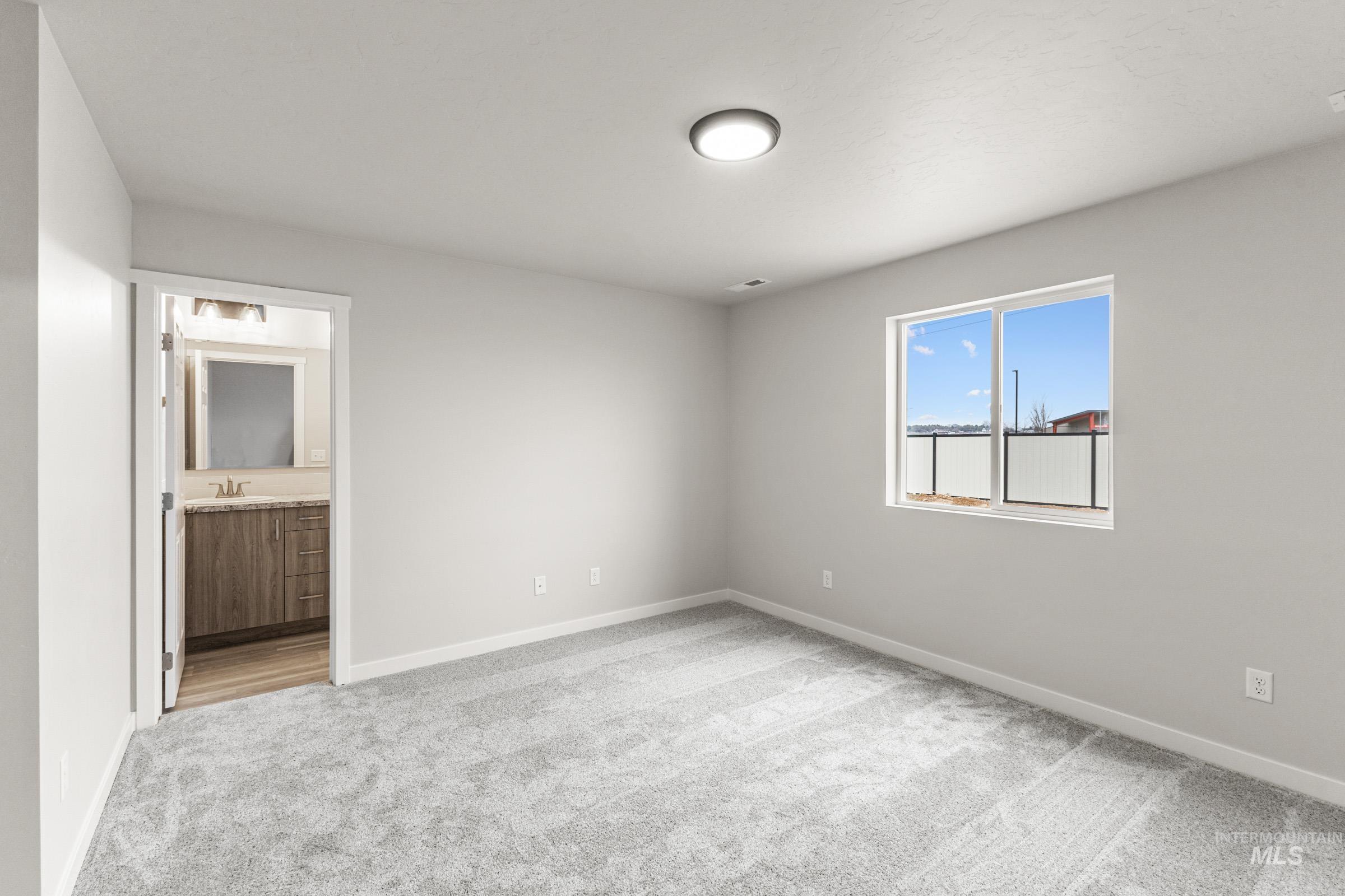Light gray carpeted room featuring a white-framed window, flush-mount ceiling light, and an entry to a bathroom with wood-finish cabinetry and a vanity mirror