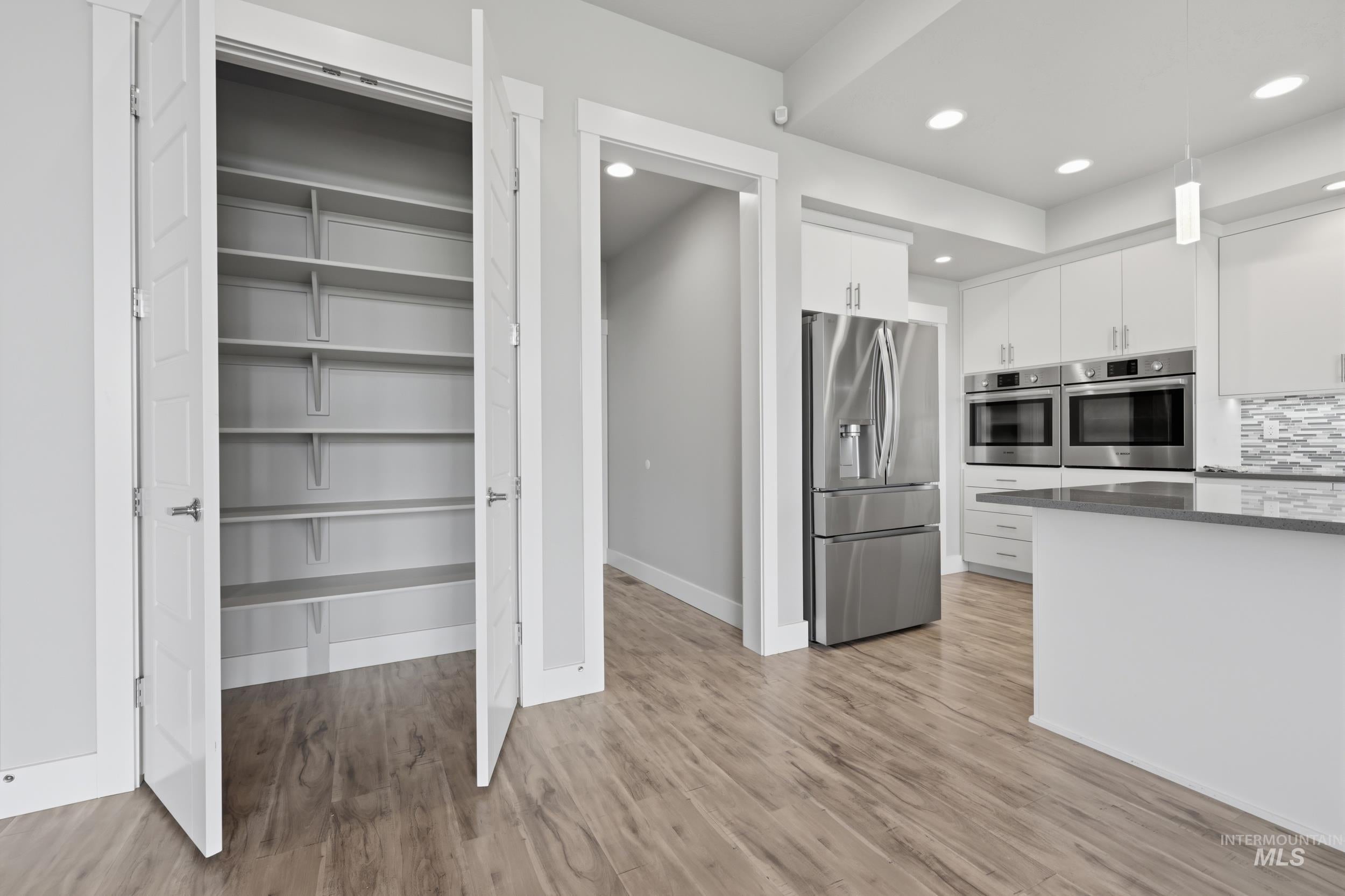 Integrated kitchen pantry with bi-fold doors, wood-finish flooring, recessed lighting, and stainless steel appliances including a refrigerator and double wall ovens