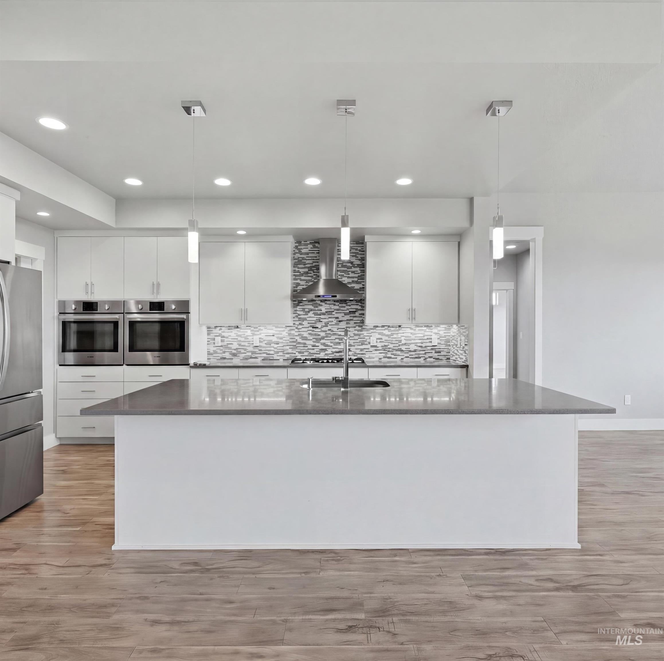 Modern kitchen featuring a large central island with a solid surface countertop, wood-finish flooring, white cabinetry, stainless steel appliances, and a mosaic tile backsplash