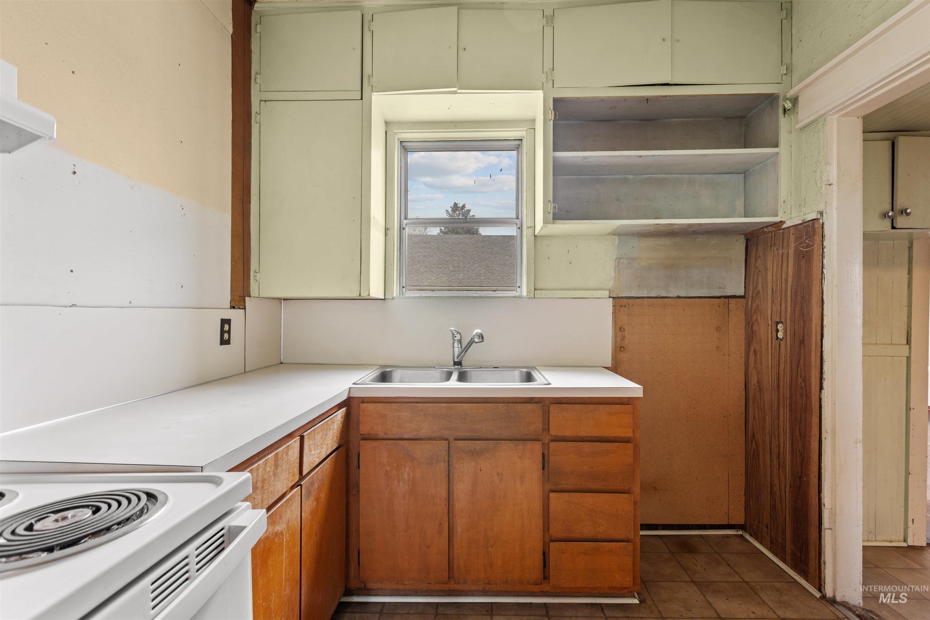 Kitchen featuring a double basin stainless steel sink, wood-finish base cabinetry, a window providing natural light, white countertops, and a white electric range