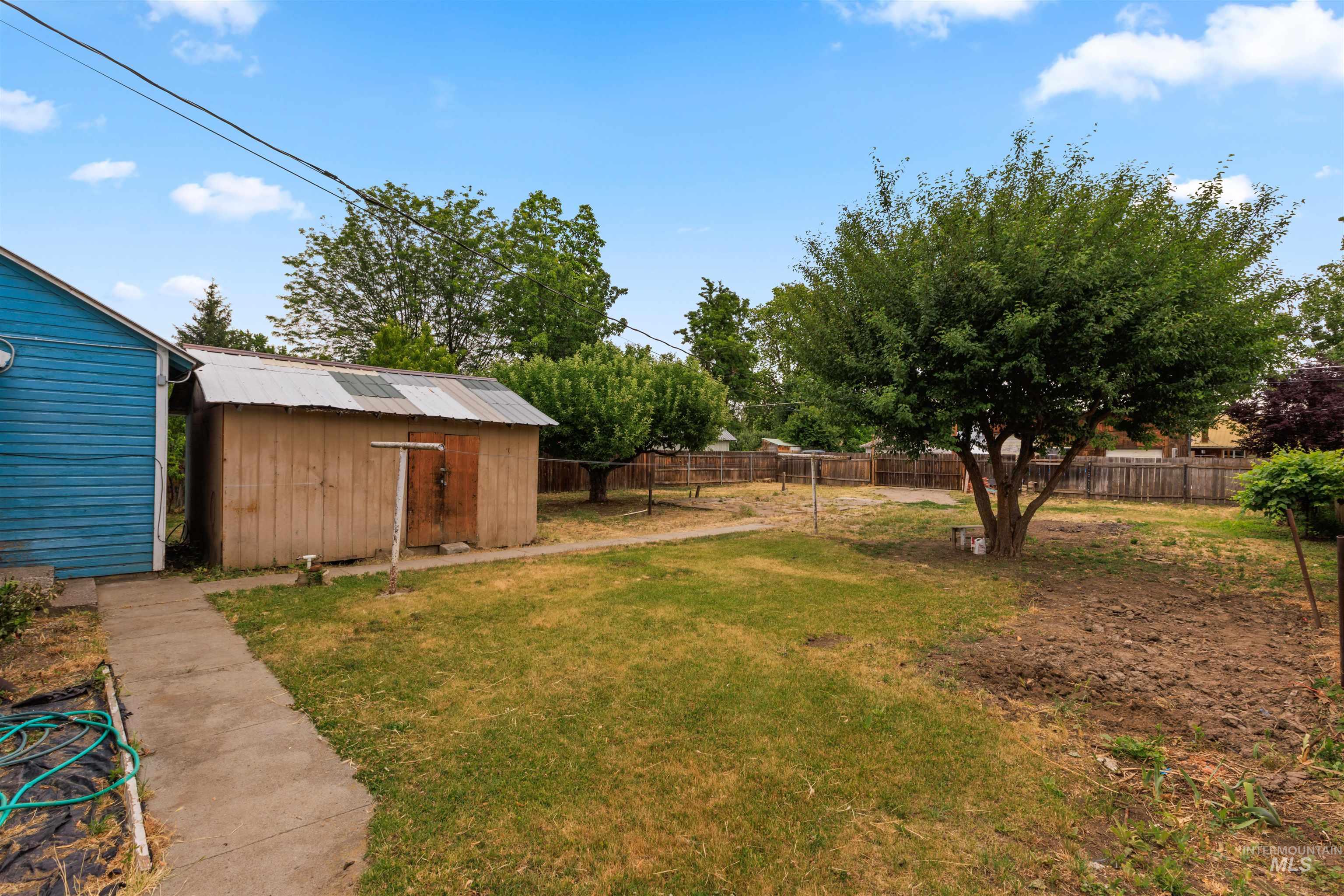 Expansive backyard featuring a mature shade tree, a wooden storage shed with a corrugated metal roof, a concrete pathway, and perimeter fencing