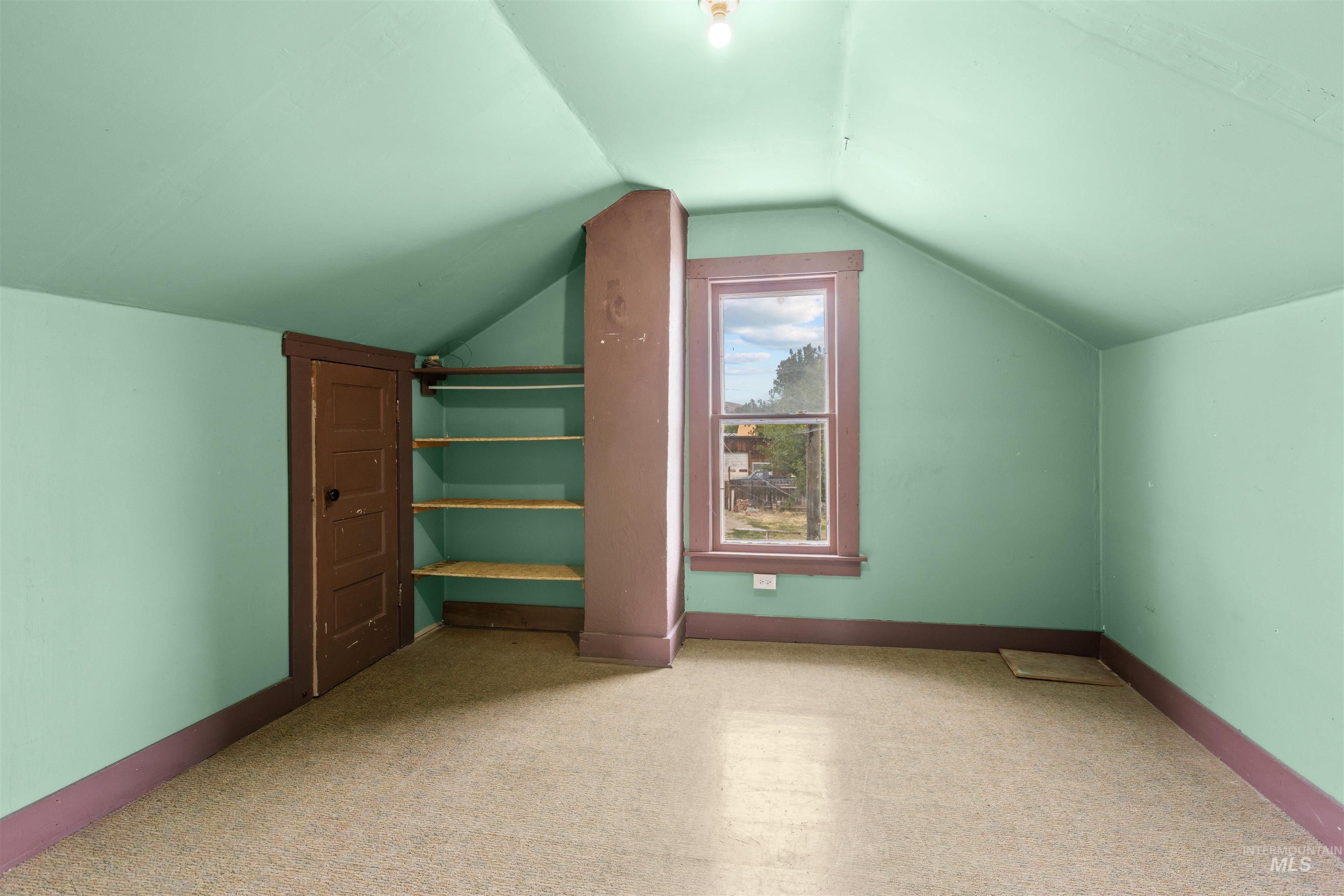 Sloped ceiling room featuring a single-pane window with wood trim, built-in shelving, a wood panel door, and light-toned carpeting
