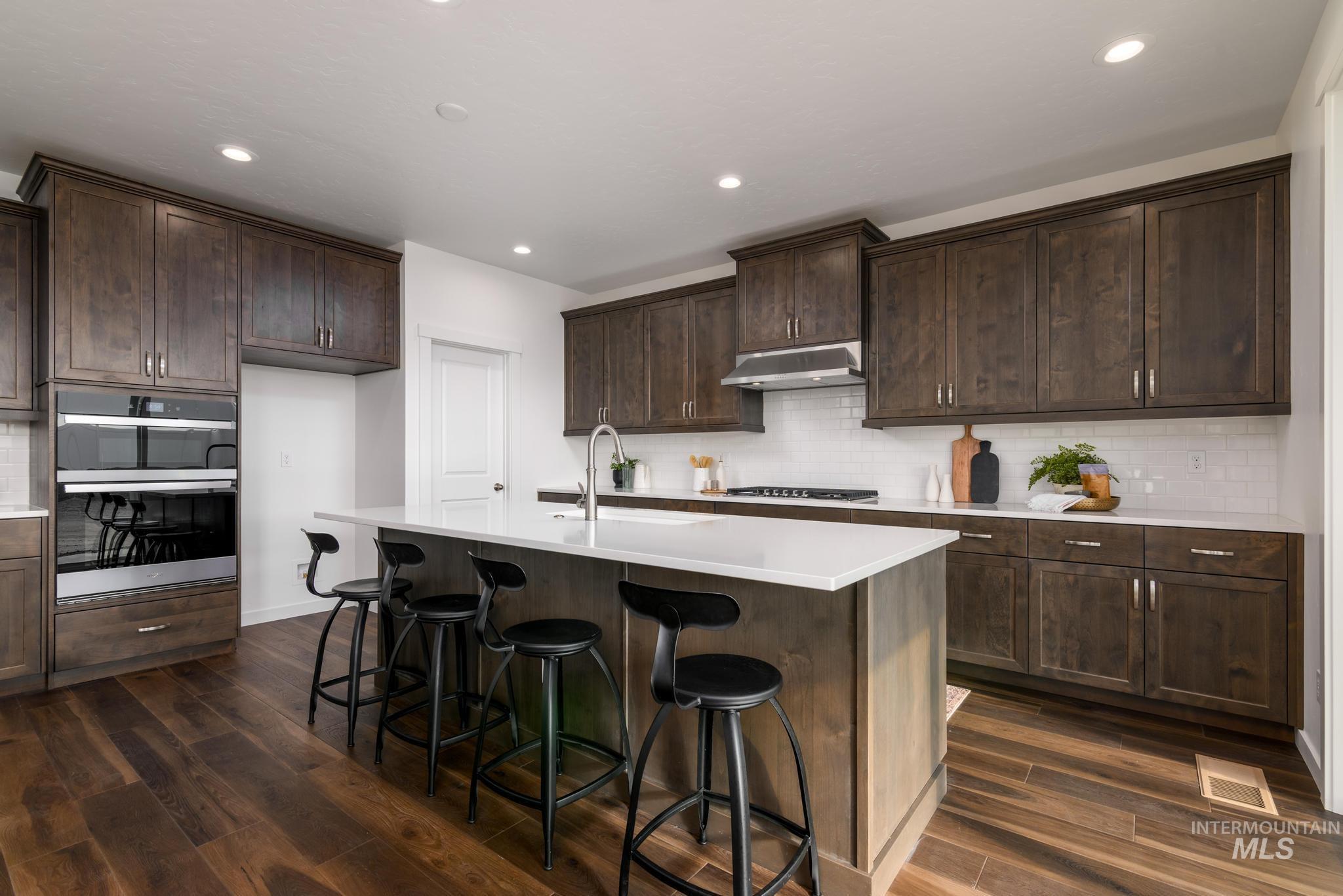 Kitchen featuring decorative backsplash, dark wood finished floors, double oven, dark brown cabinets, and a kitchen bar