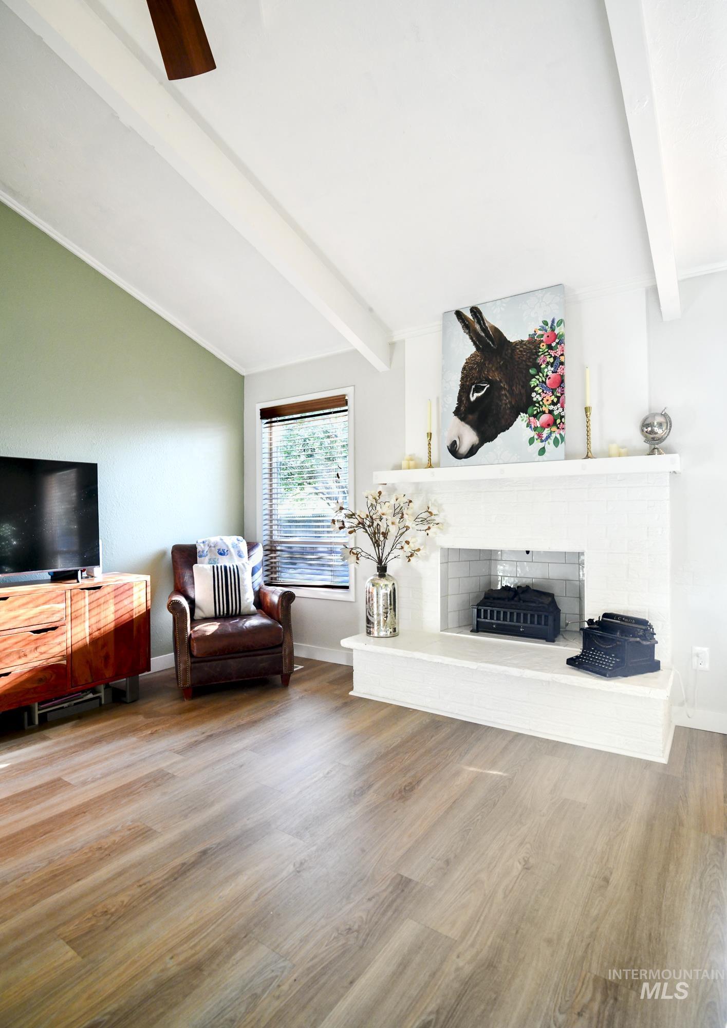 Living area featuring a brick fireplace, light wood finished floors, ceiling fan, and vaulted ceiling with beams