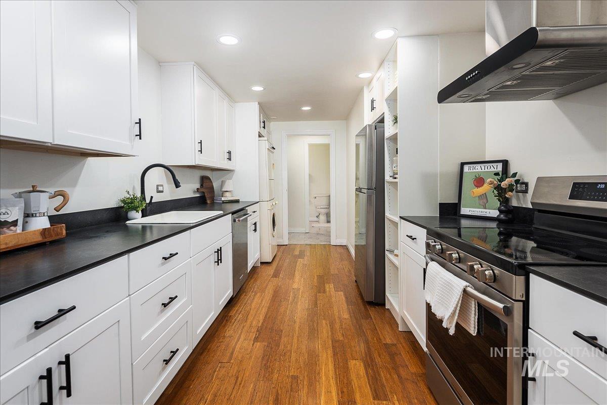 Kitchen featuring dark countertops, stainless steel appliances, wall chimney range hood, white cabinetry, and recessed lighting
